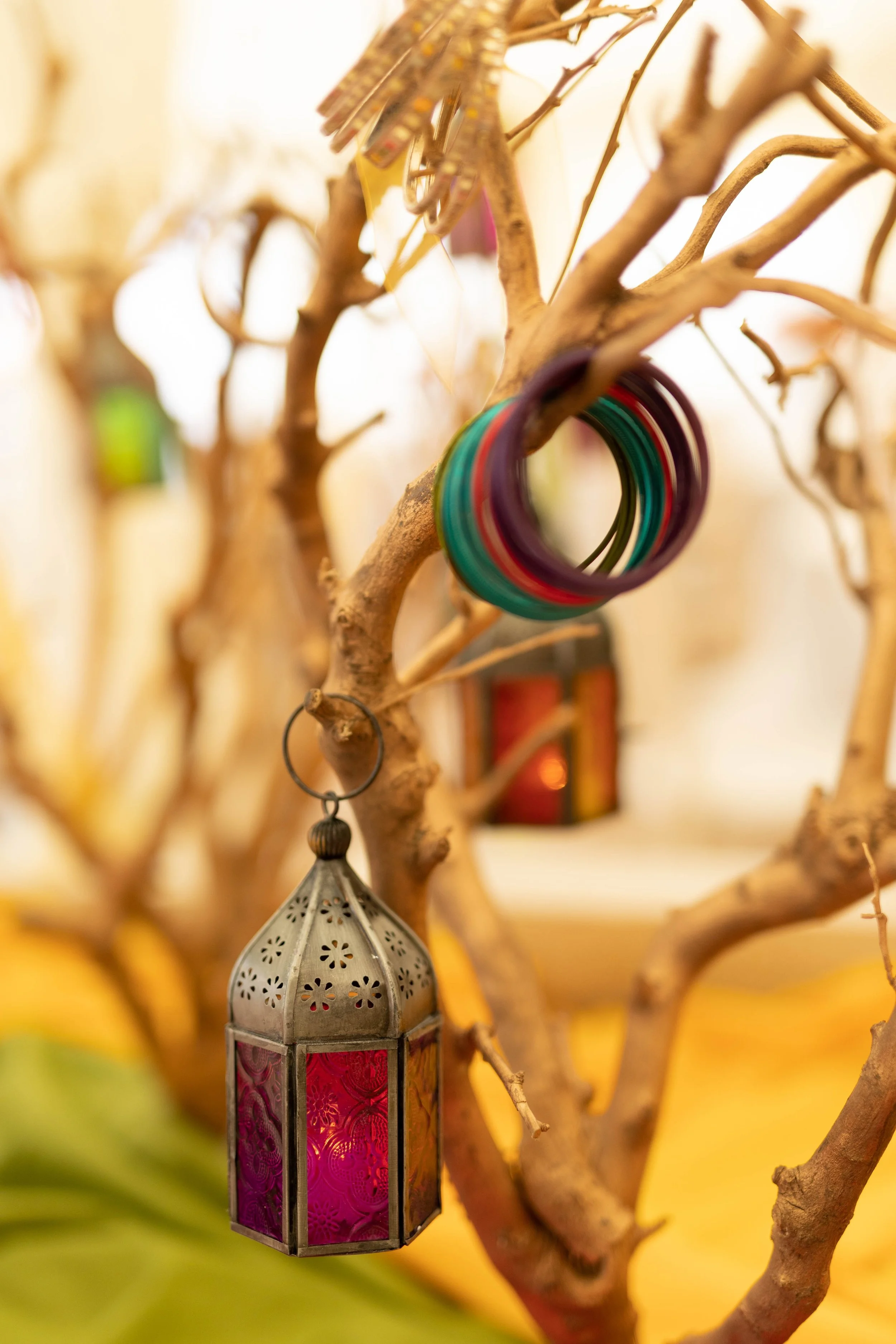A dried, leafless decorative tree with colorful bangles hanging from its branches and a metal lantern with red and purple glass panels hanging from one of the branches.