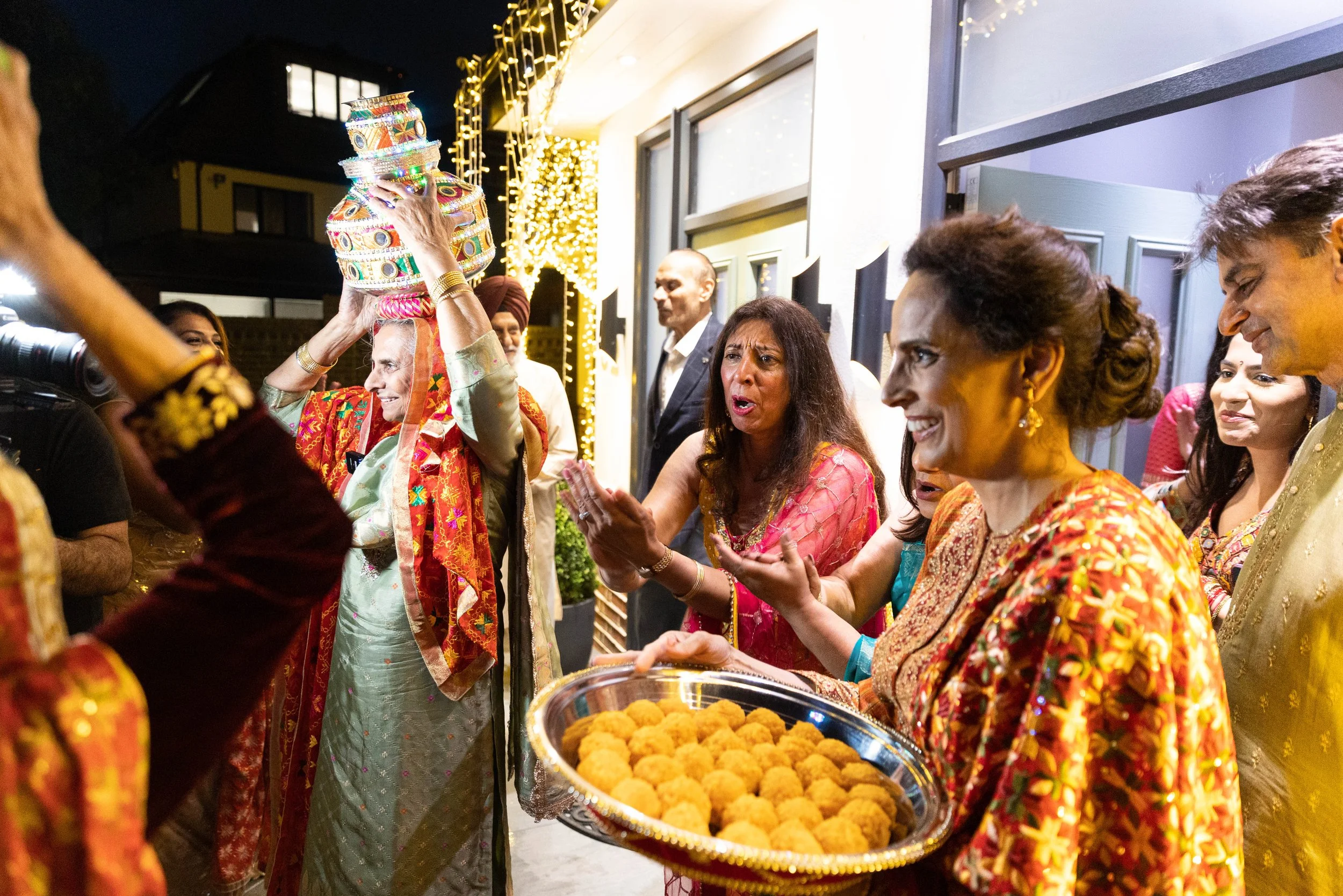 An Indian woman in traditional attire holds a large silver tray of Indian sweets. She is surrounded by women in colorful sarees and men in traditional clothing, participating in a festive celebration at night outside a house decorated with lights.