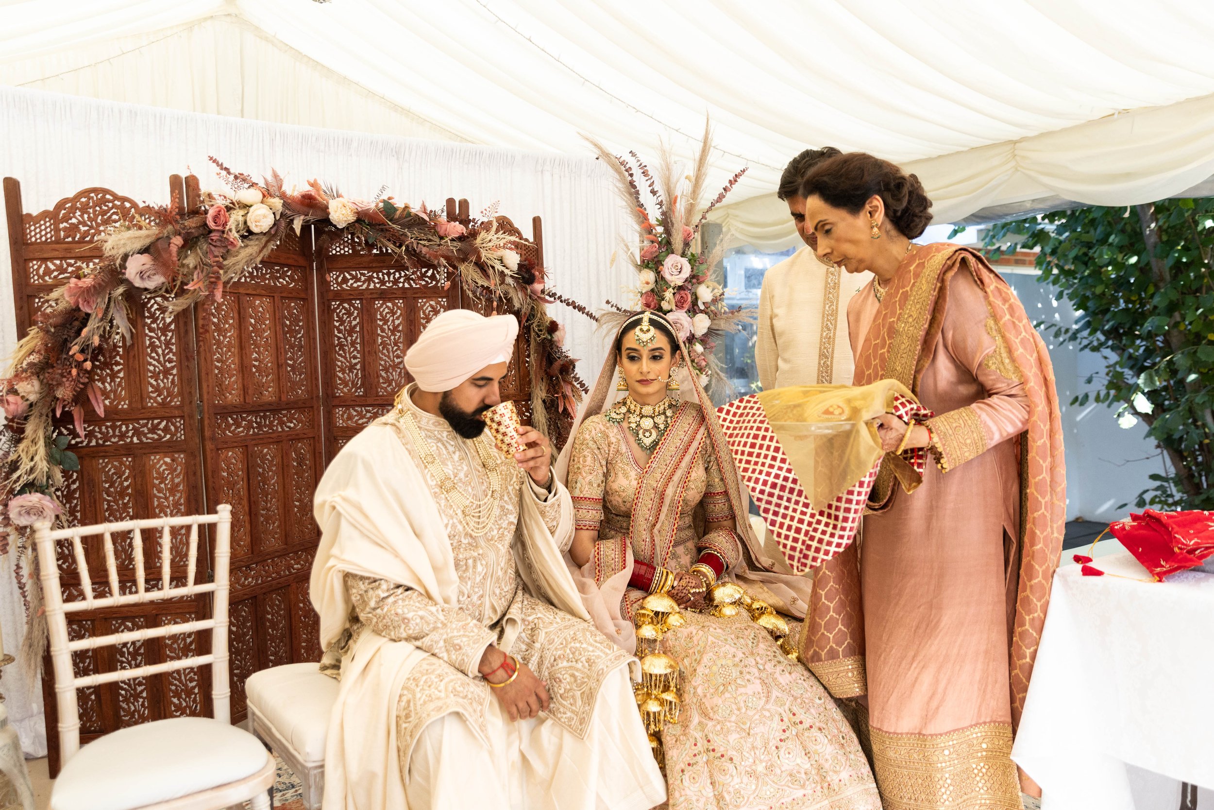 Indian wedding ceremony with a bride and groom dressed in traditional attire, seated with family members in a decorated tent, floral decorations, and a woman holding a gift.