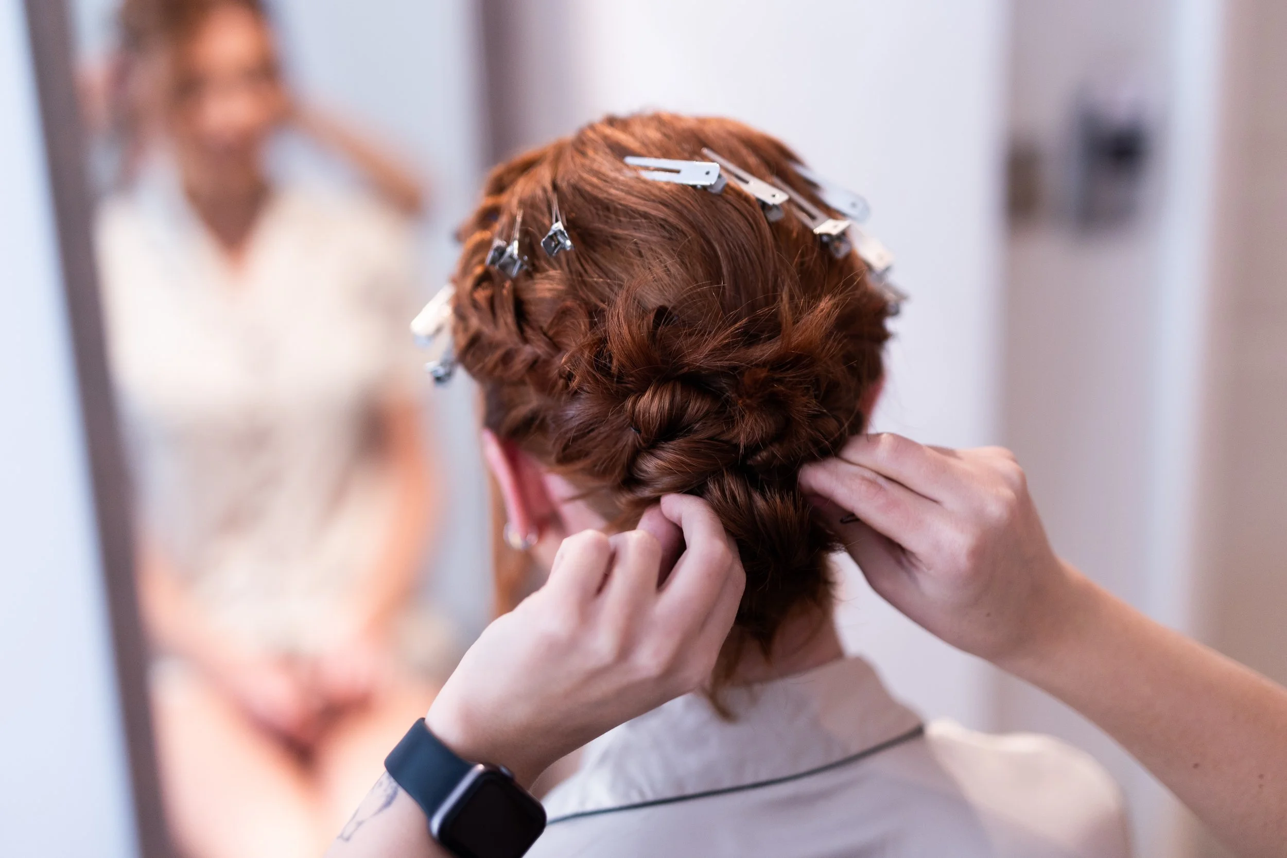 A woman with red hair is getting her hair styled with curlers and braids, looking into a mirror. Another woman is smiling in the background.