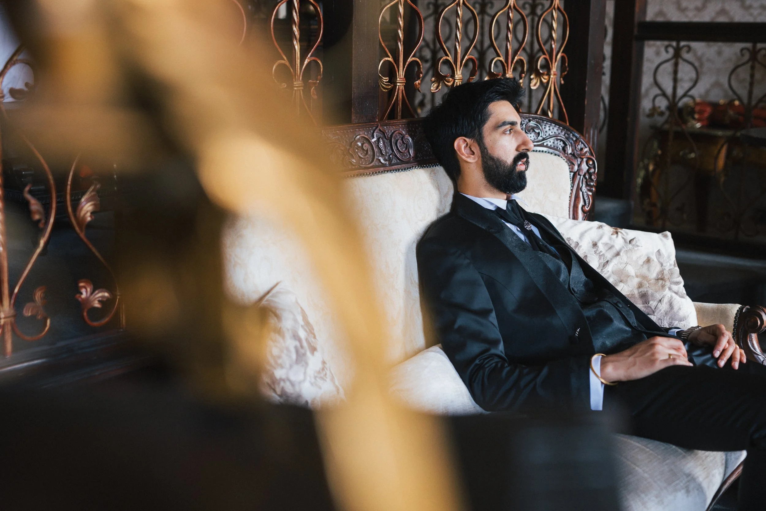 A man with a beard wearing a black tuxedo sitting on an ornate vintage sofa, looking to the right, in a luxurious room with decorative ironwork and patterned cushions.