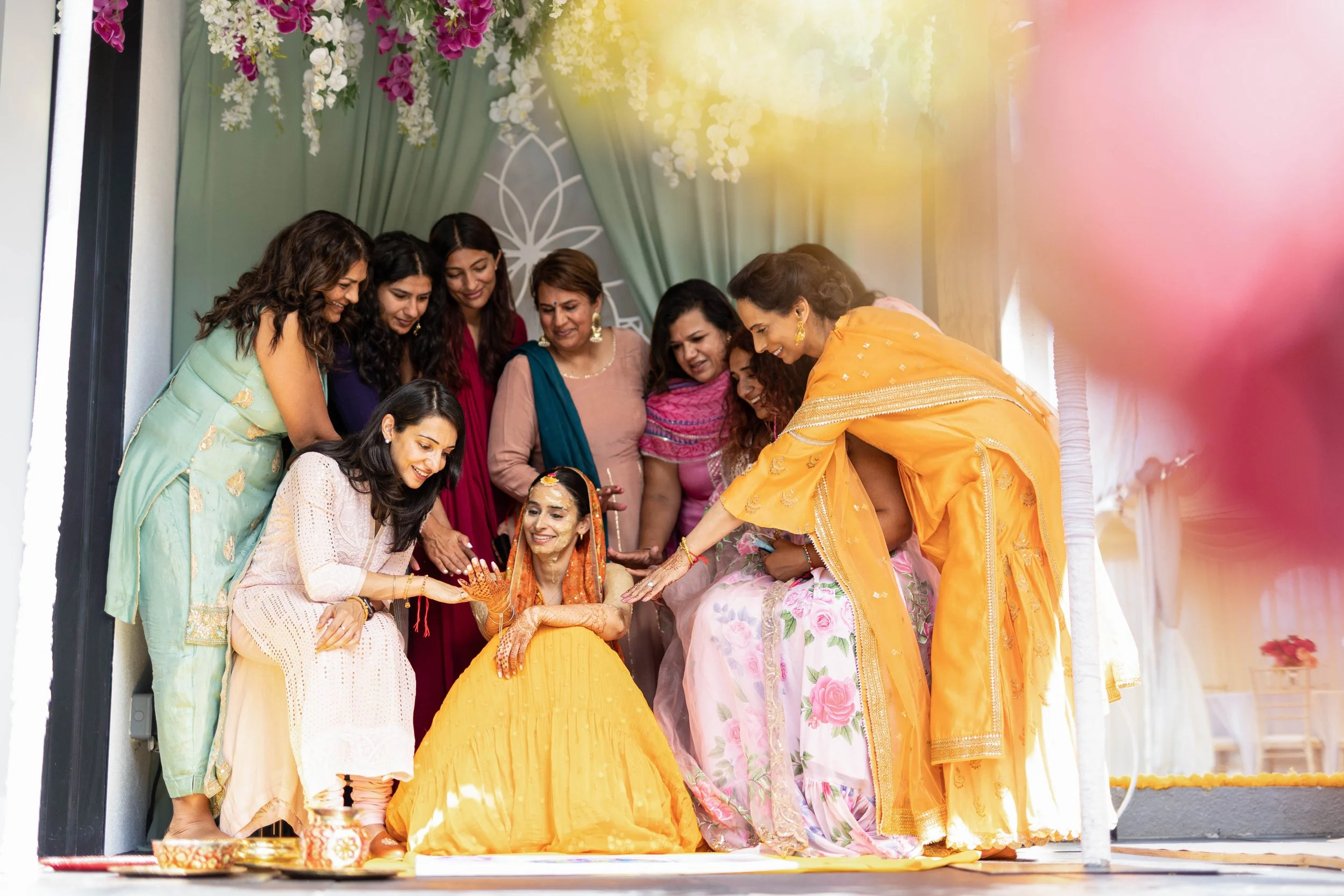 Group of women celebrating a traditional Indian wedding, with the bride sitting in the center dressed in a yellow and orange sari, surrounded by friends and family, some touching her hands and smiling.