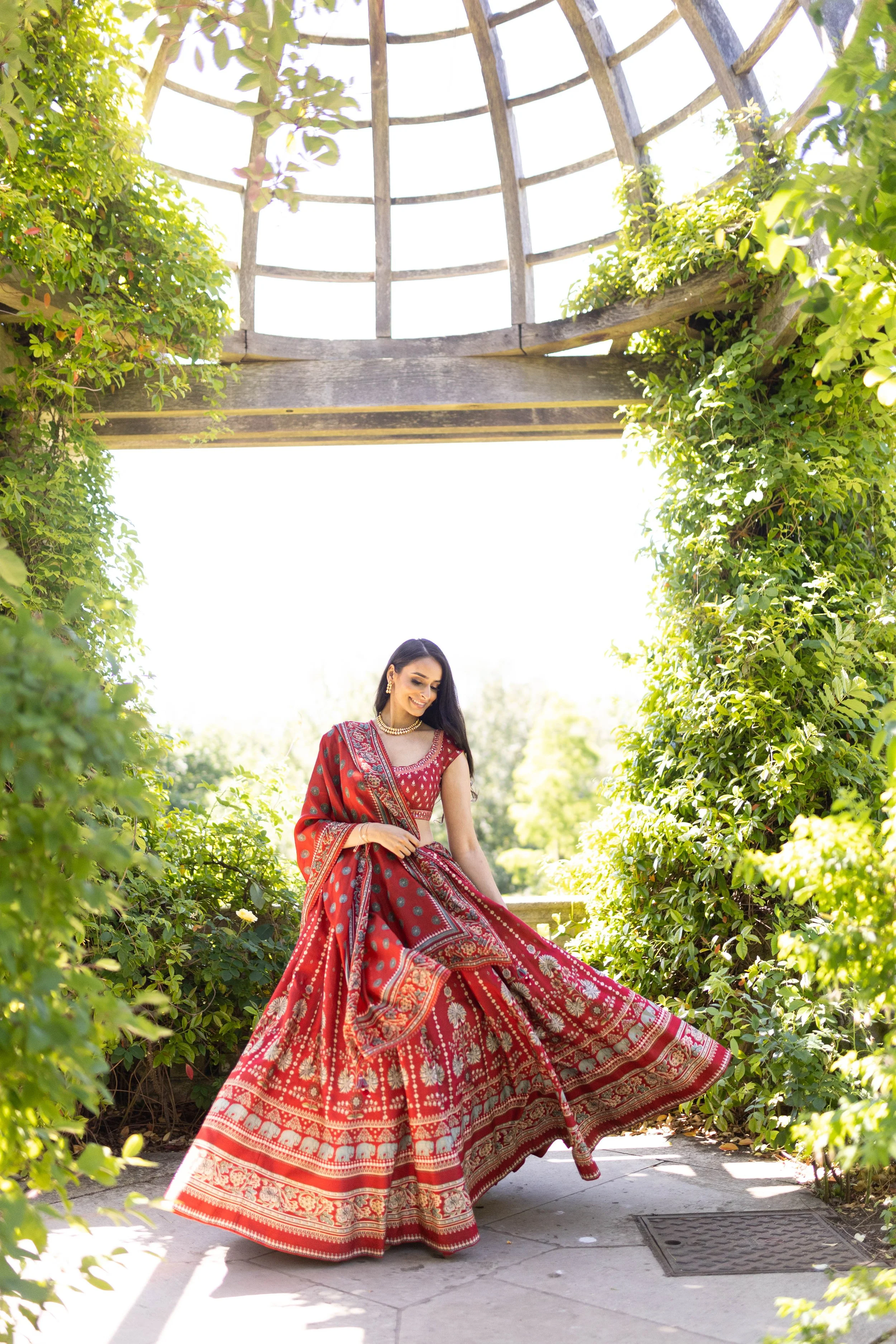 A woman in a red traditional Indian dress with gold embroidery is smiling and posing in a garden under a wooden archway surrounded by green foliage on a bright sunny day.