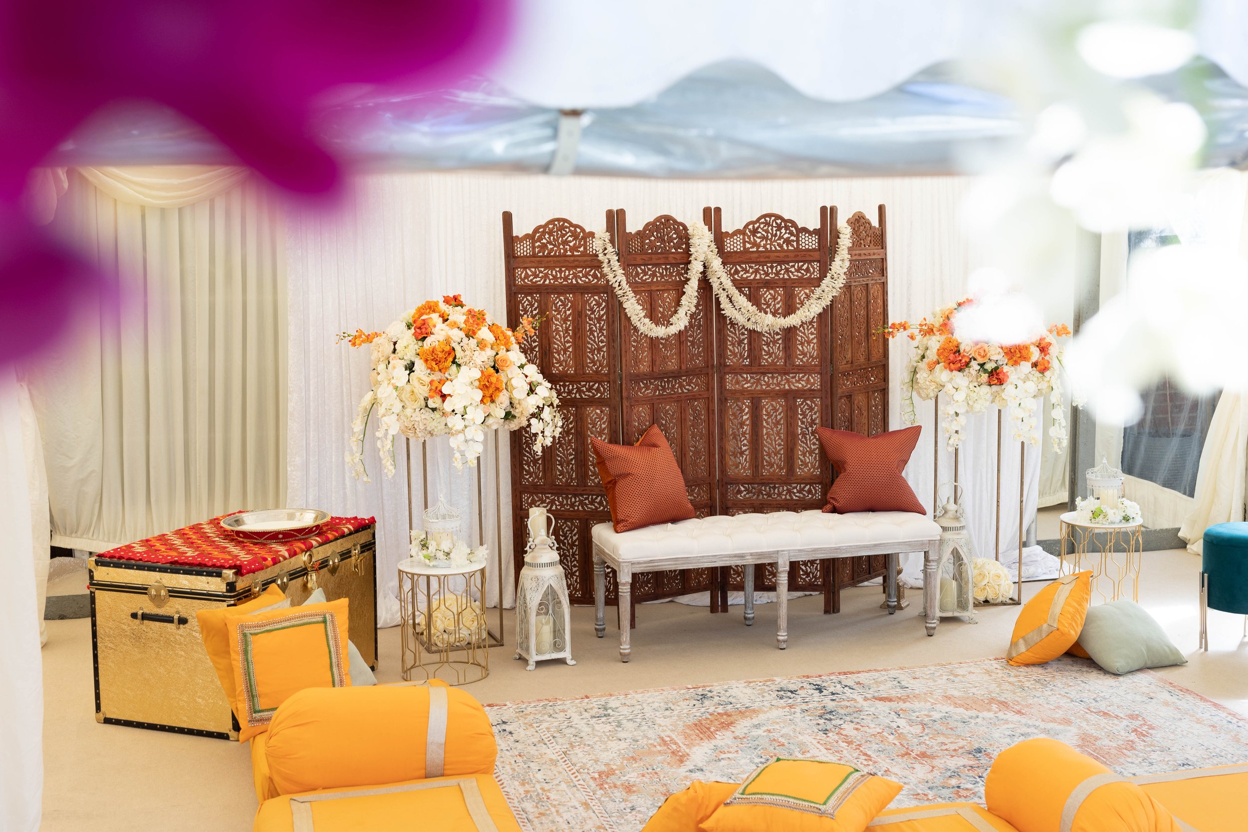 A decorated wedding or event setting with a wooden screen, floral arrangements, pillows, and cushions in warm tones, arranged in a cozy, elegant manner.