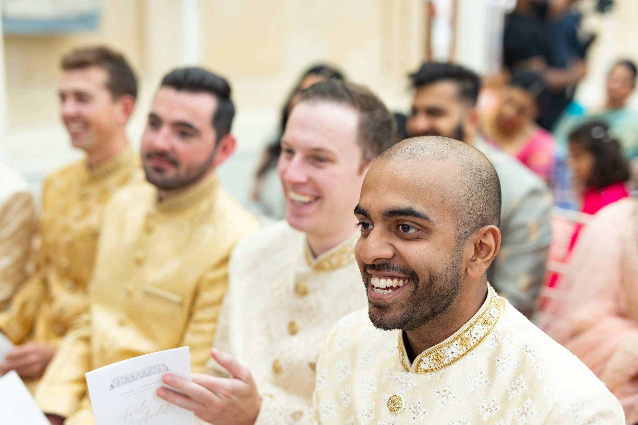 Men dressed in traditional Indian attire attending a cultural or religious ceremony, sitting in a row and smiling.