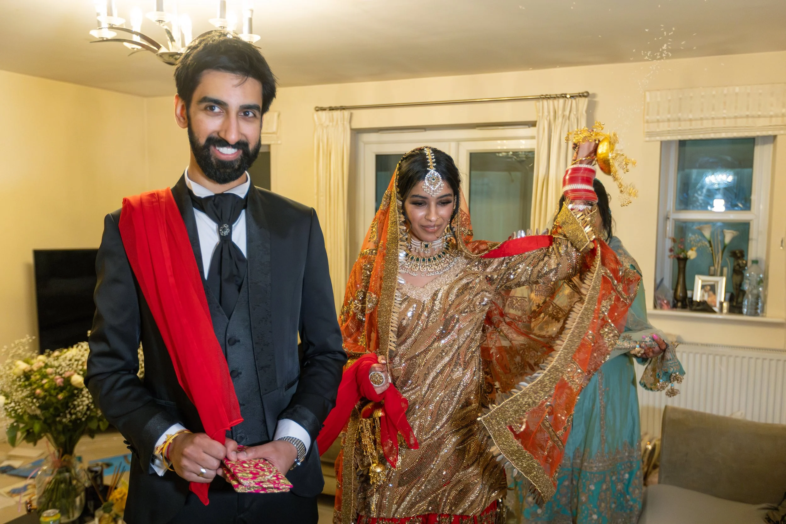 A couple dressed in wedding attire, the groom in a black suit with a red stole, smiling at the camera, and the bride in a traditional gold and red Indian outfit with jewelry, lifting her arm in a dance pose inside a decorated room.