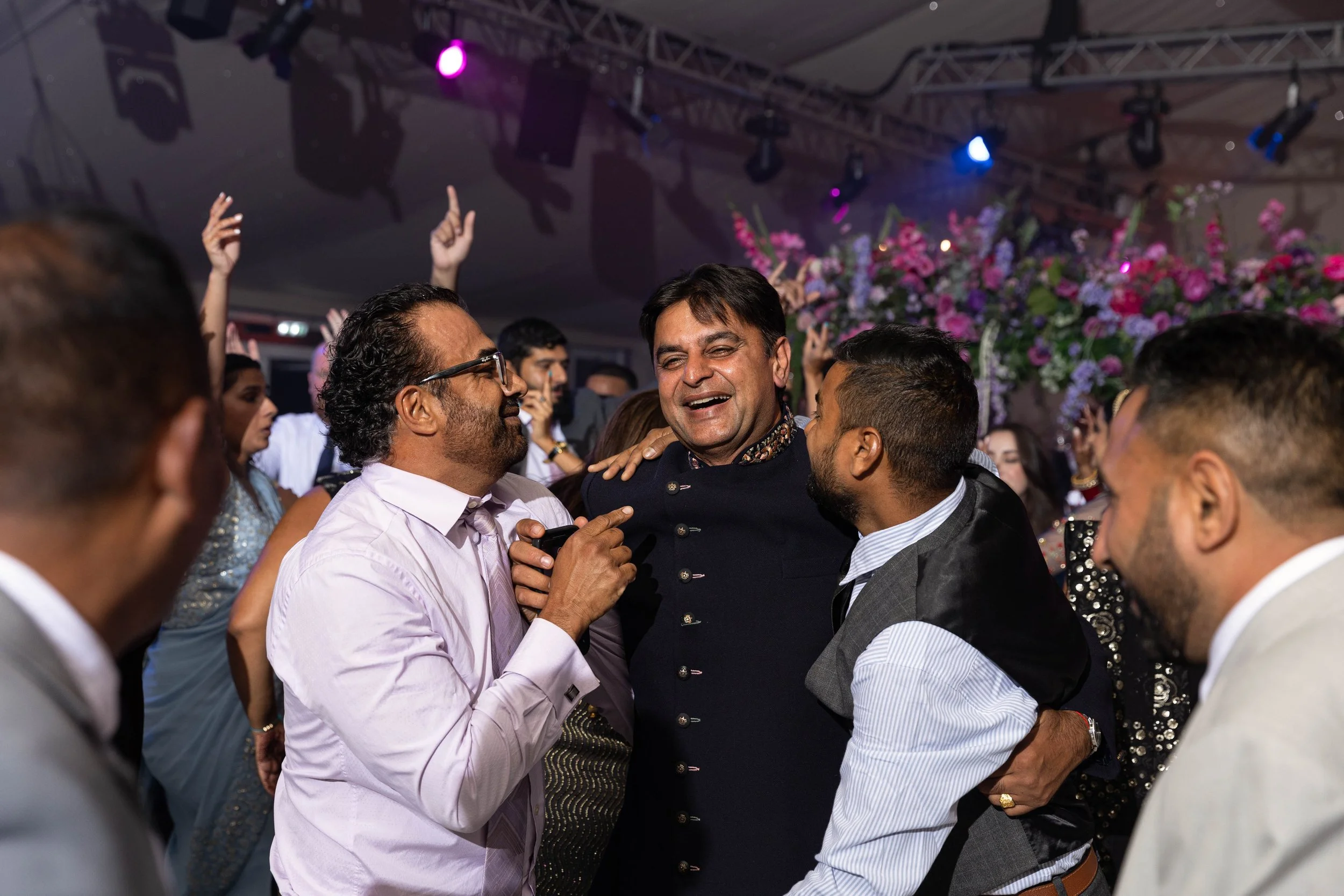 People dancing and celebrating at a wedding reception with pink and purple floral decorations and colorful stage lighting.