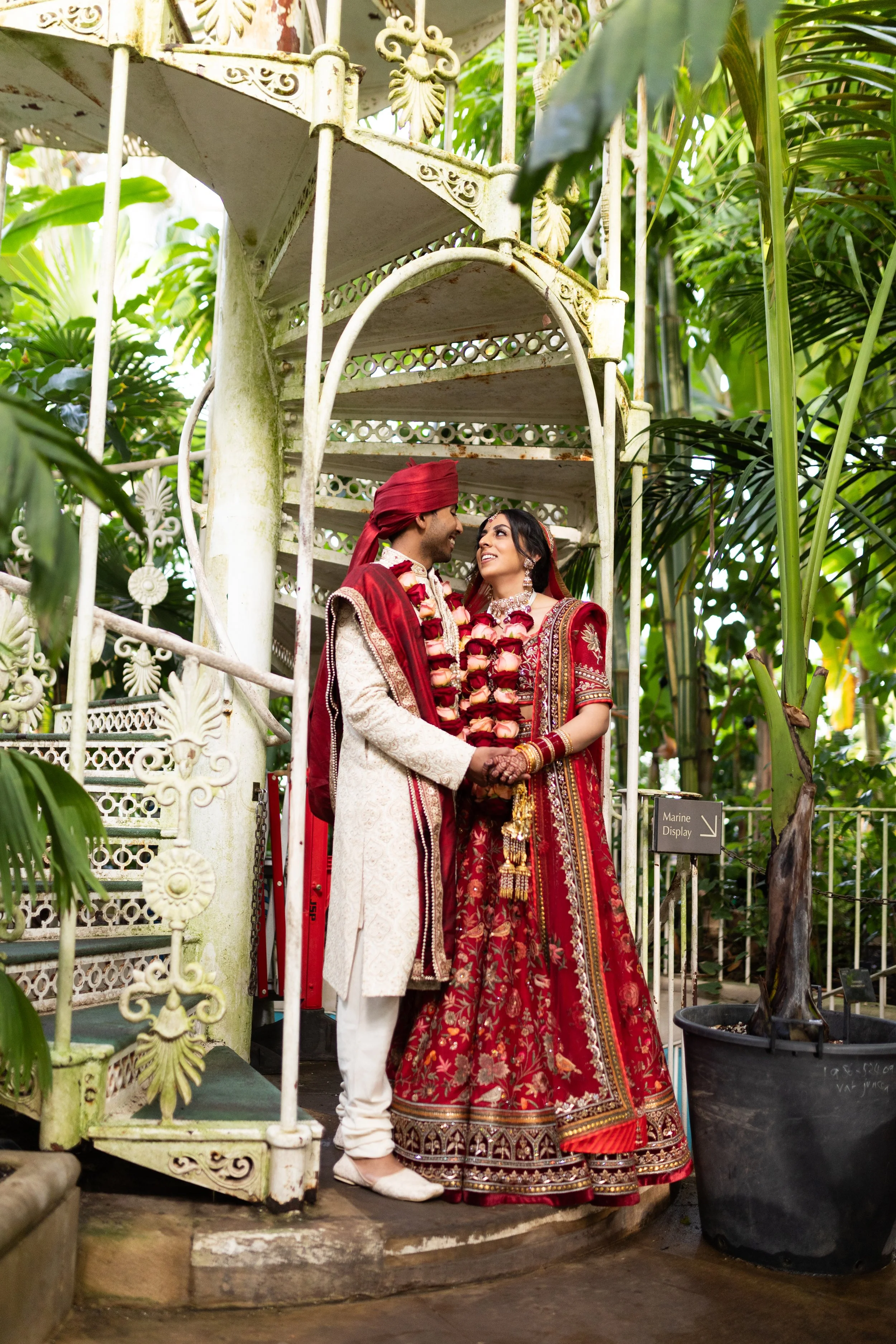 A newlywed couple dressed in traditional Indian wedding attire, holding hands and smiling at each other underneath a vintage spiral staircase surrounded by lush greenery.