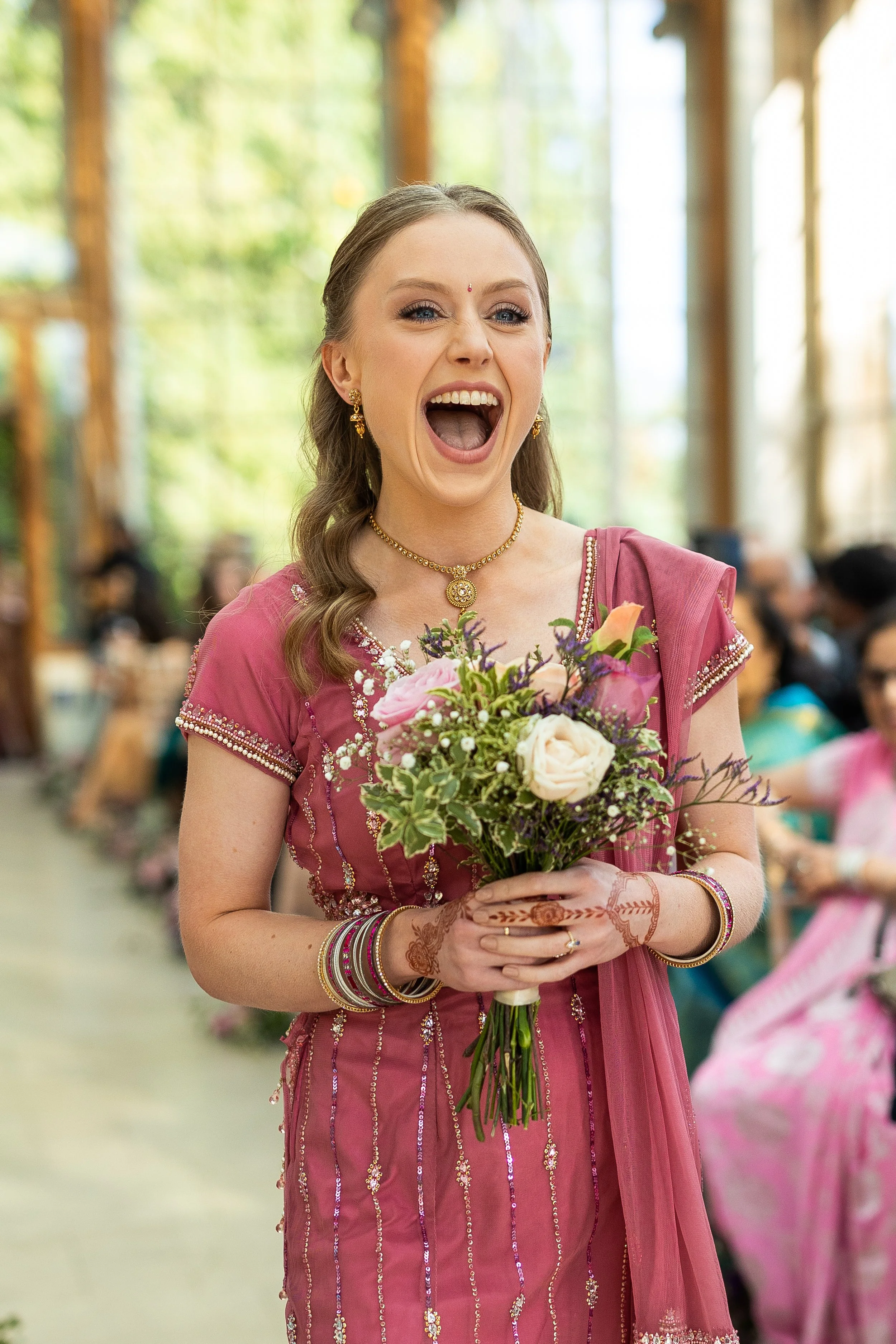 A woman wearing a pink traditional outfit holding a bouquet of flowers at a celebration, possibly a wedding, with other guests seated in a bright, indoor space.