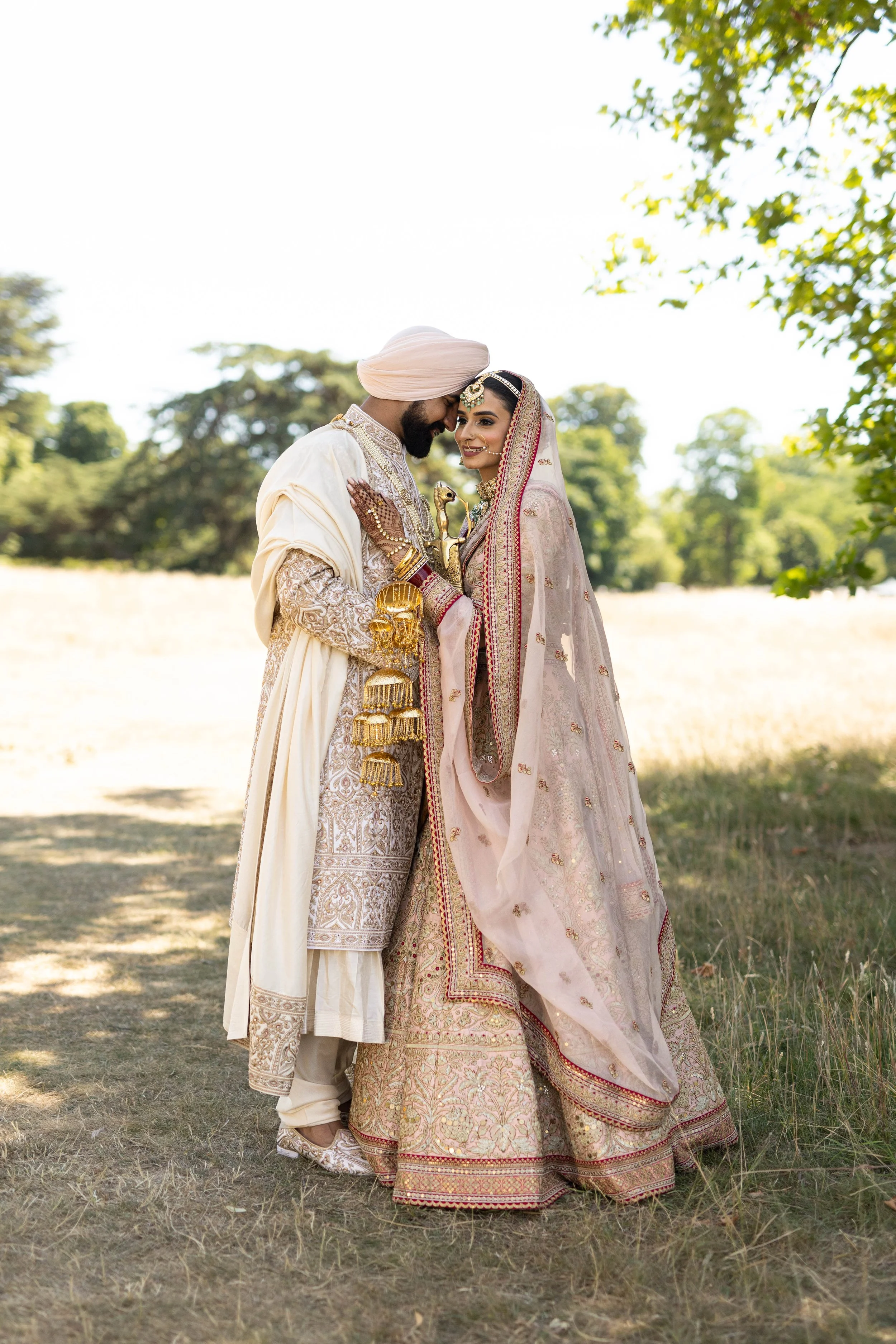 A couple dressed in traditional Indian wedding attire standing closely outdoors in a grassy area with trees in the background, sharing an intimate moment.
