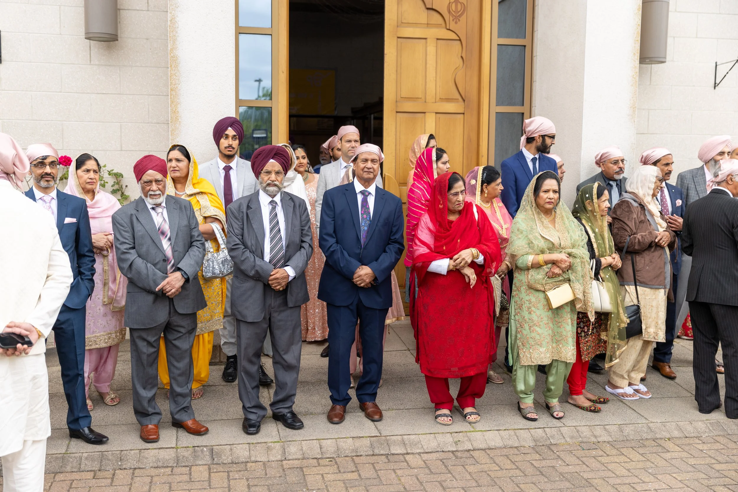 Group of Indian people, mostly wearing traditional clothes and turbans, standing in front of a church entrance, attending a wedding or celebration.
