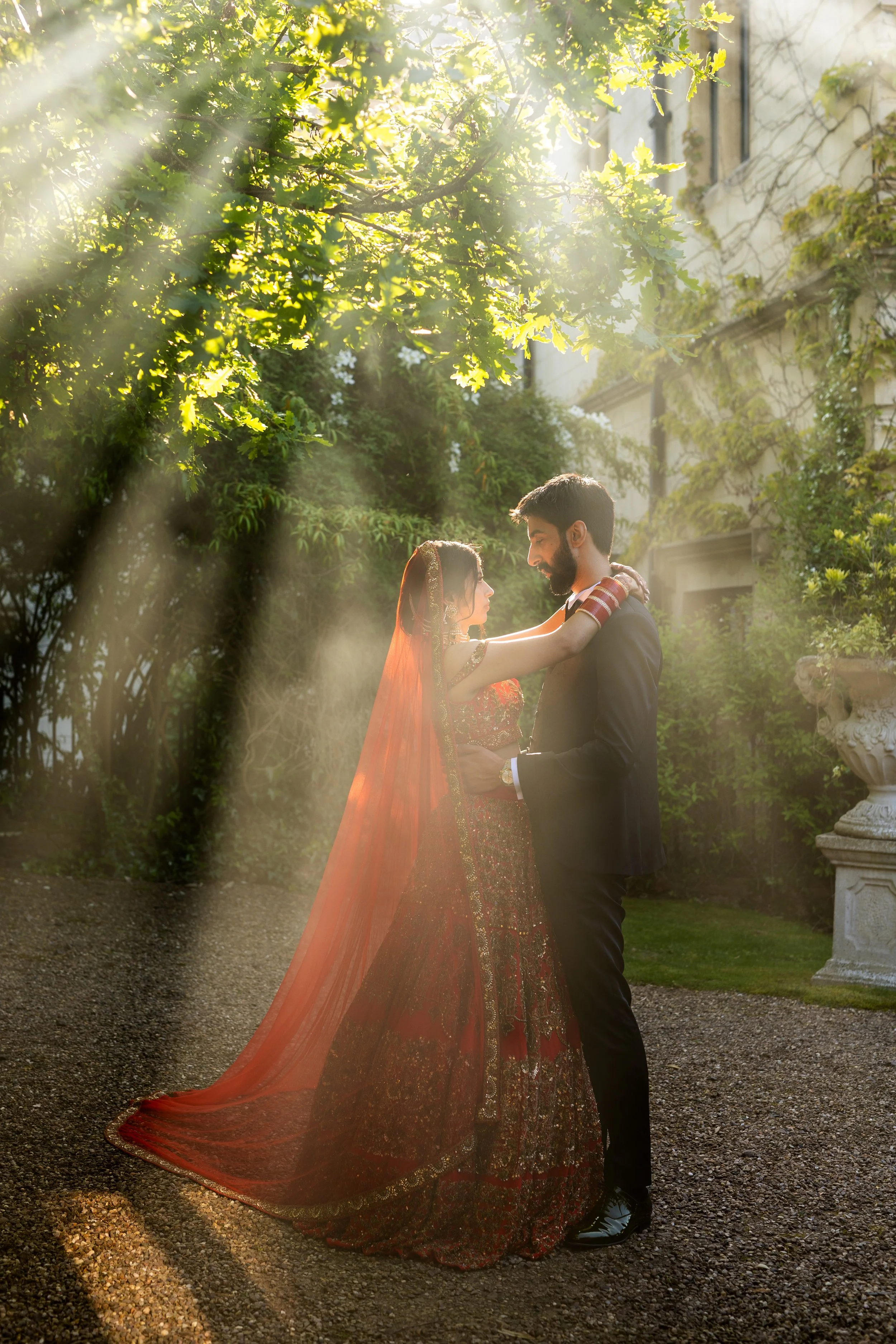 A couple in traditional Indian wedding attire embracing outdoors in sunlight surrounded by greenery.