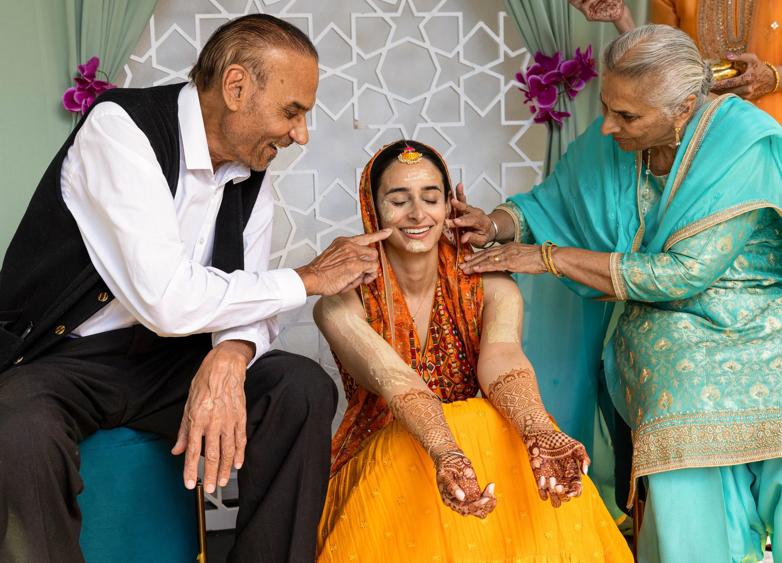A young woman in traditional Indian attire receives a pre-wedding ceremony involving turmeric application from two elders, a man and a woman, who are lovingly and smilingly applying the turmeric to her face and arms during a festive celebration.