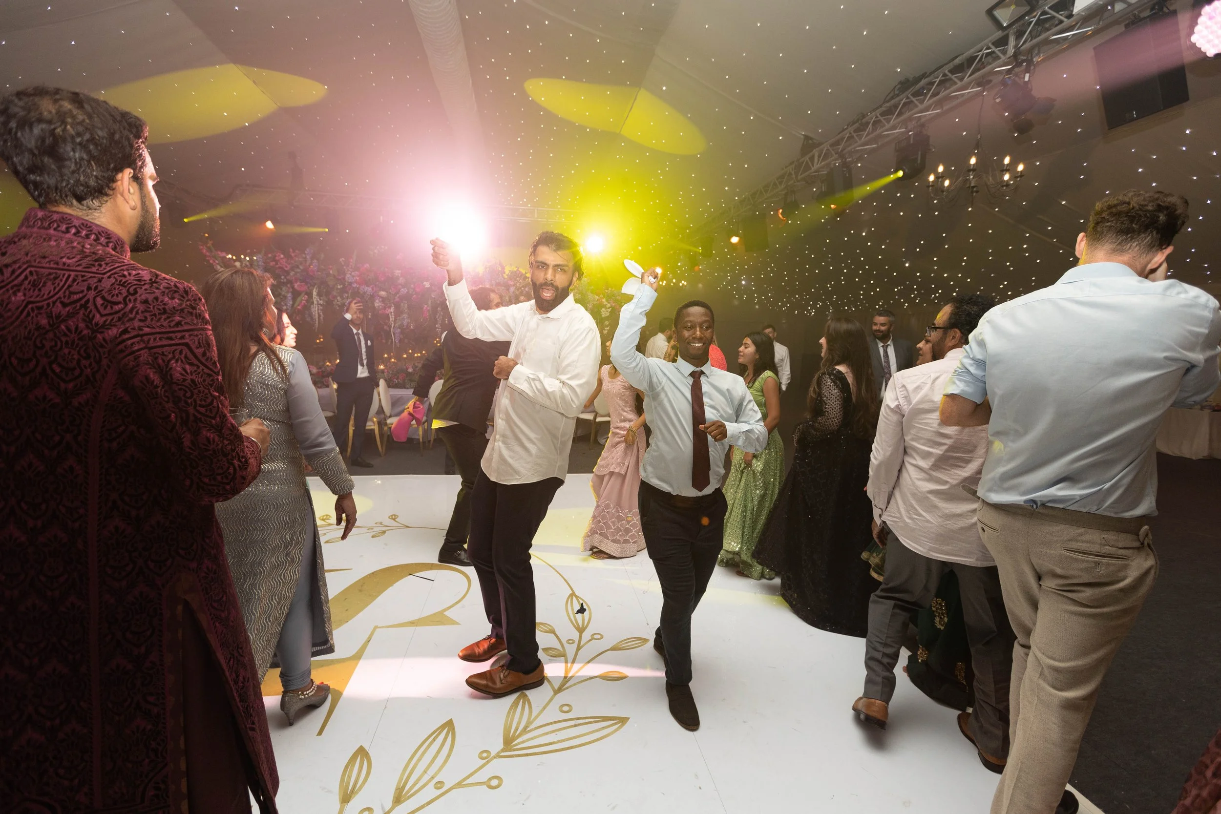 People dancing and celebrating at a lively party or wedding reception with colorful lighting and decorated venue.
