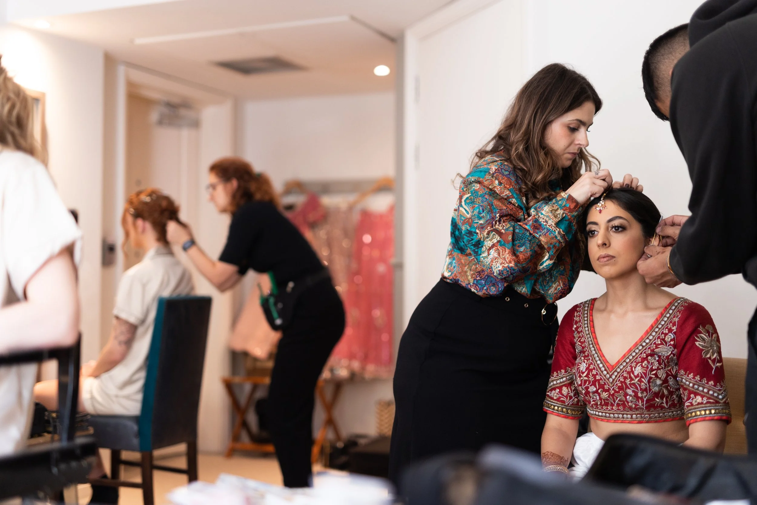 Women preparing models for a traditional Indian dance performance, with one woman applying jewelry to a seated woman dressed in a red and gold costume.