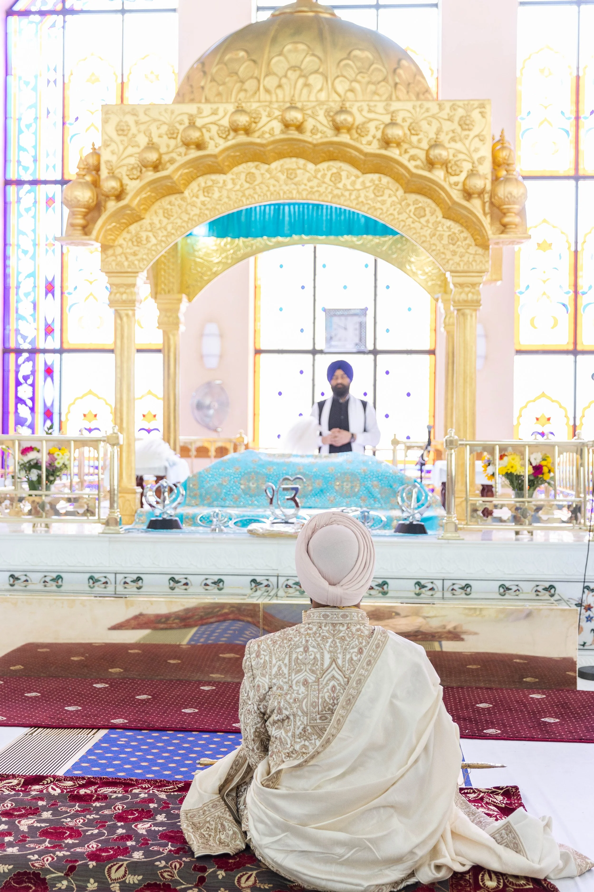 A man in traditional Sikh attire kneeling in prayer at a temple, facing the altar with a priest standing behind it. The temple features colorful stained glass windows and intricate gold and white decorations.