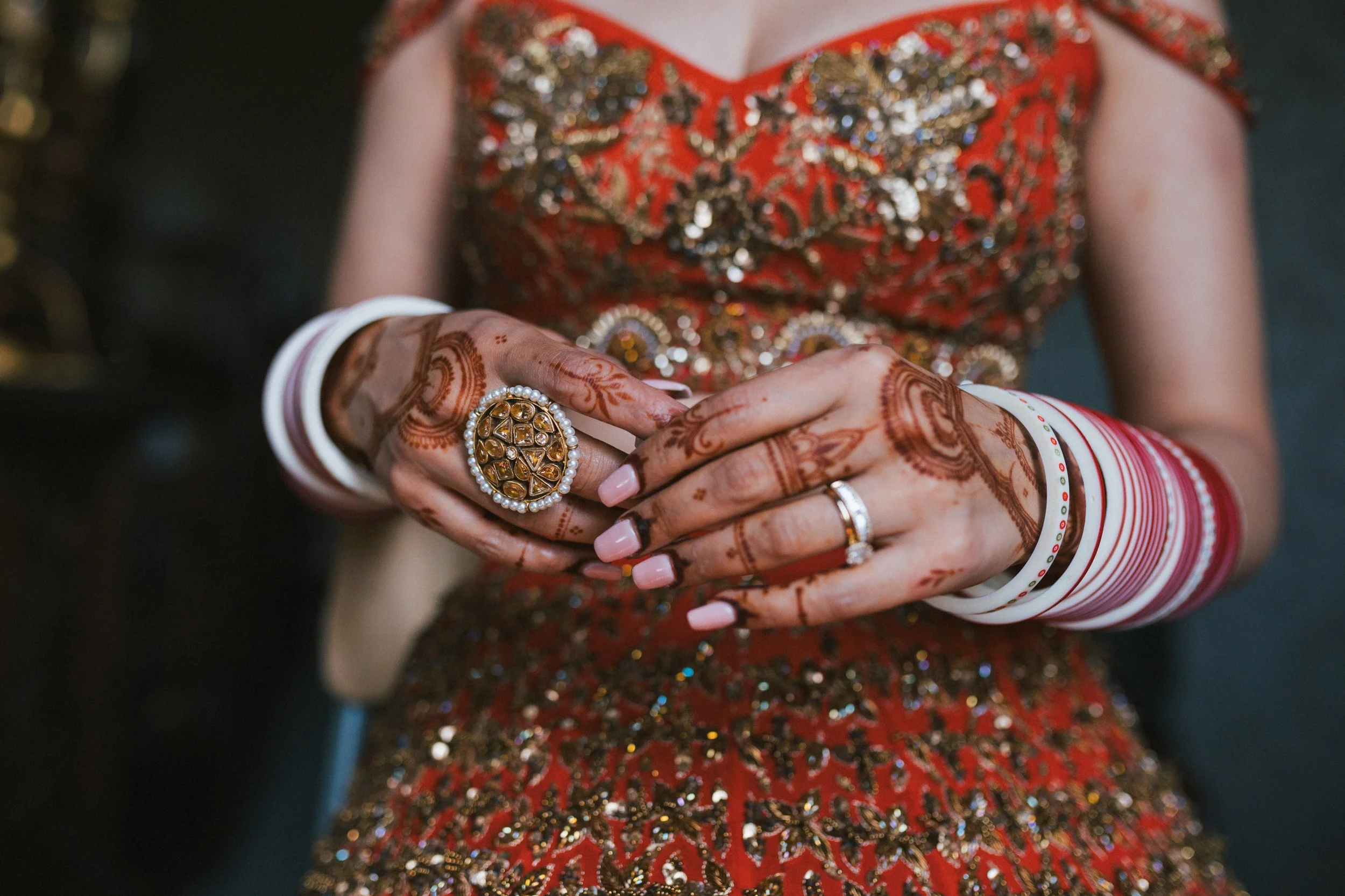 Close-up of a woman wearing traditional red and gold embroidered attire, with henna designs on her hands, adorned with rings, bangles, and a large statement ring with gemstones, capturing a moment at a celebration or wedding.