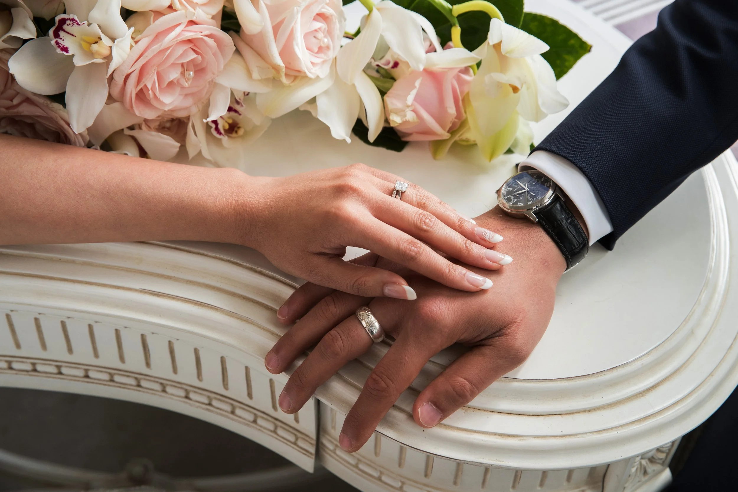 A newlywed couple's hands are shown with the bride's hand resting on top of the groom's hand, both wearing wedding rings. The bride's hand has a large diamond engagement ring and a wedding band, and her nails are manicured. The groom's left hand has a wedding band and he is wearing a wristwatch with a black leather strap. The couple's hands are on a white ornate table with a bouquet of pink and white flowers nearby.