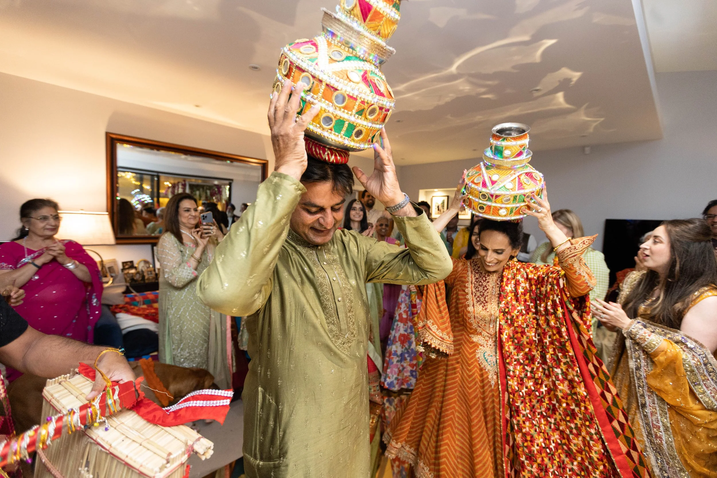 People in traditional Indian attire celebrating and performing a ritual with decorated pots on their heads at an indoor event.