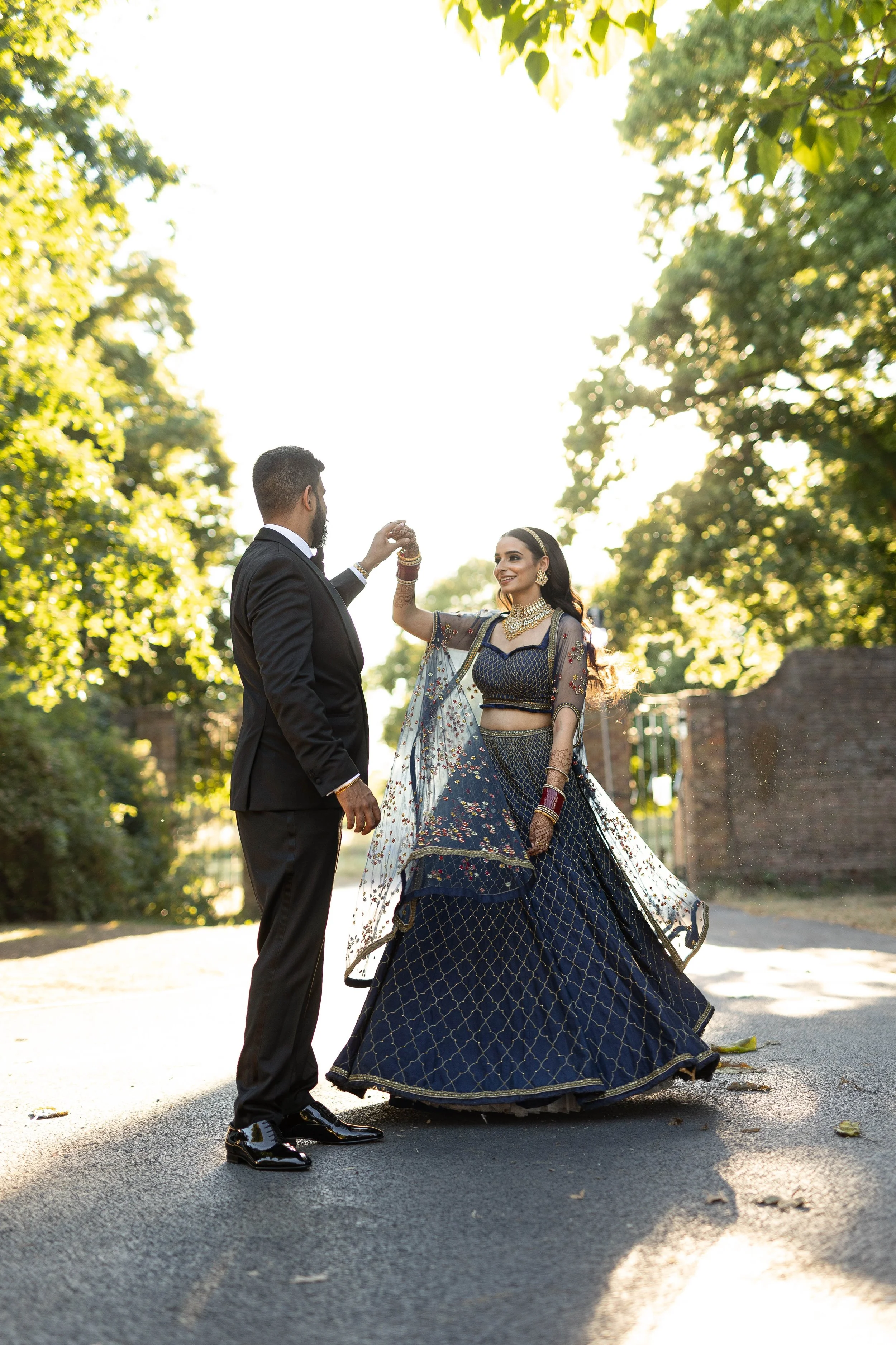 A man in a black suit and a woman in a traditional blue and gold Indian dress doing a dance outdoors on a sunny day, surrounded by green trees.