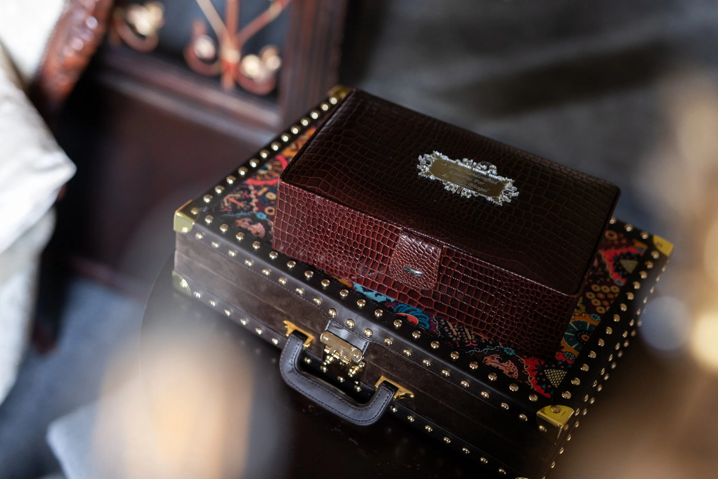 A decorative box with a floral pattern and studded border, topped with a brown box with a shiny, textured surface and a decorative metal label