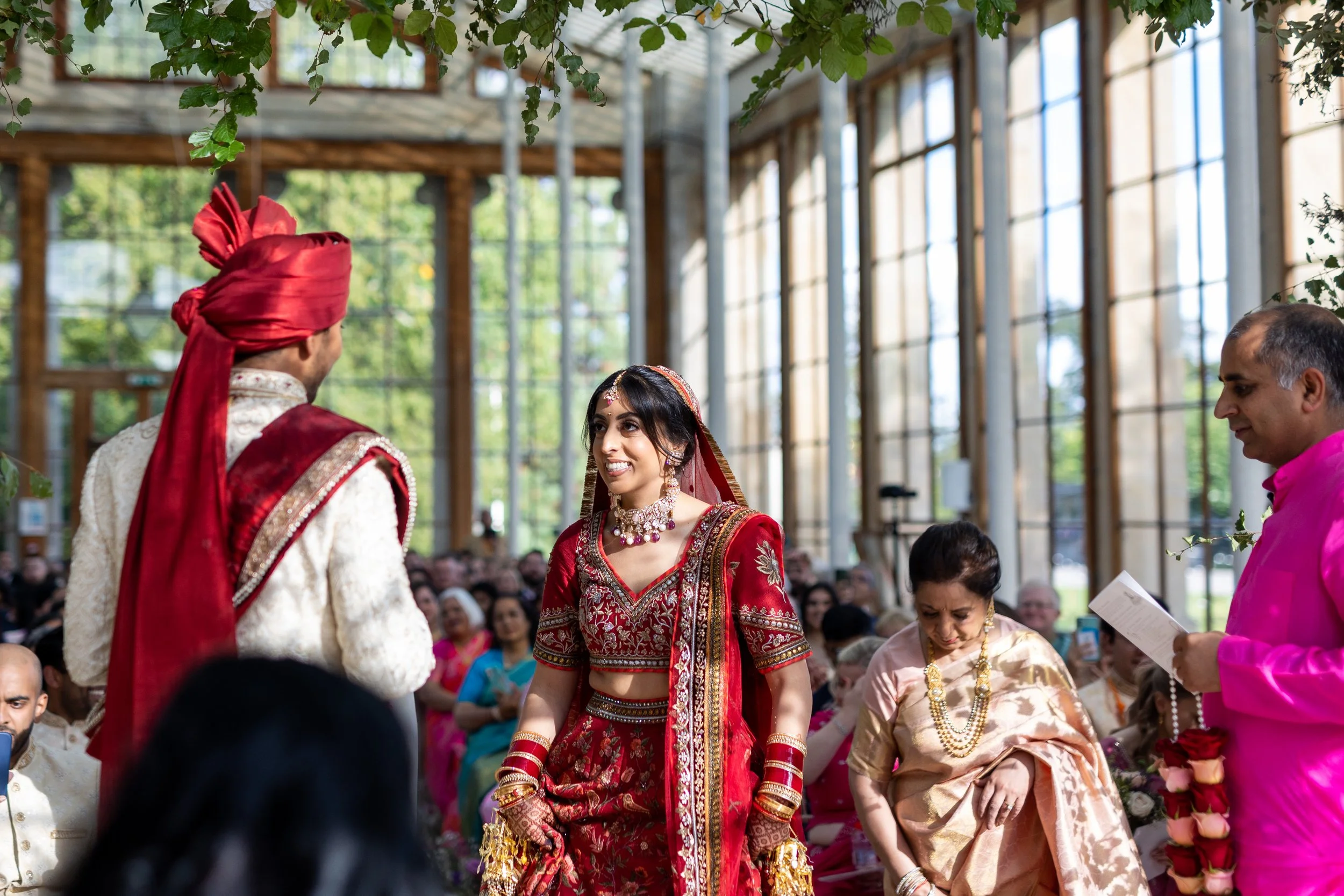 Indian wedding ceremony with a bride in red traditional attire, standing next to a groom in cream and red, inside a glass-ceiling venue filled with guests.