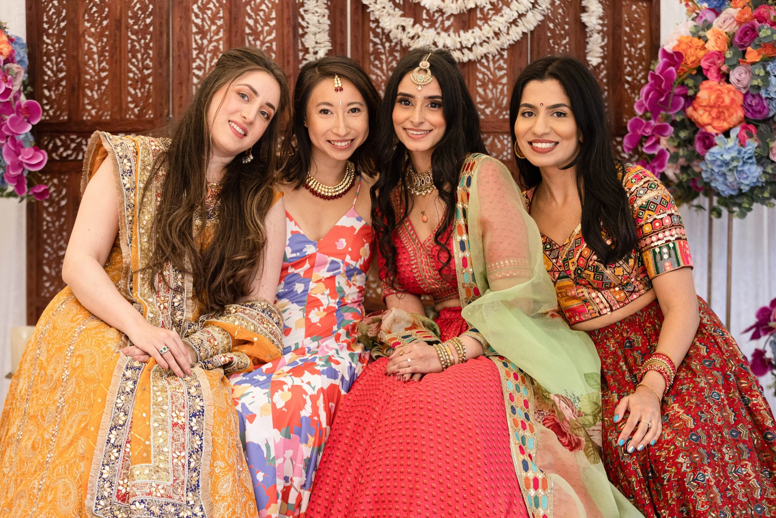 Four women dressed in colorful traditional South Asian attire smiling and sitting together at an indoor event with floral decorations.