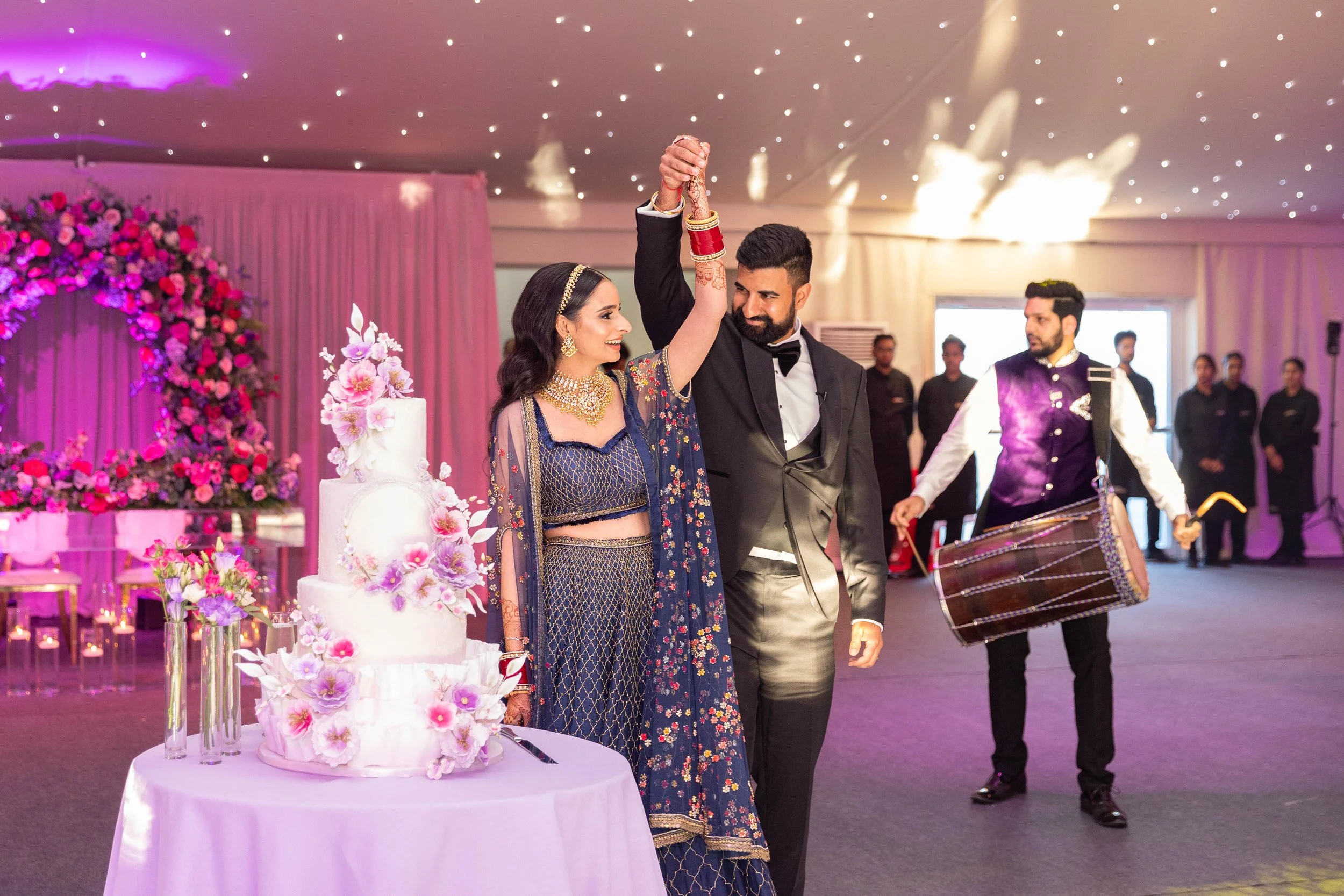 A bride and groom are dancing together at their wedding reception, holding hands above their heads. The bride is dressed in traditional Indian attire with jewelry, and the groom is in a black suit with a bow tie. A wedding cake decorated with flowers