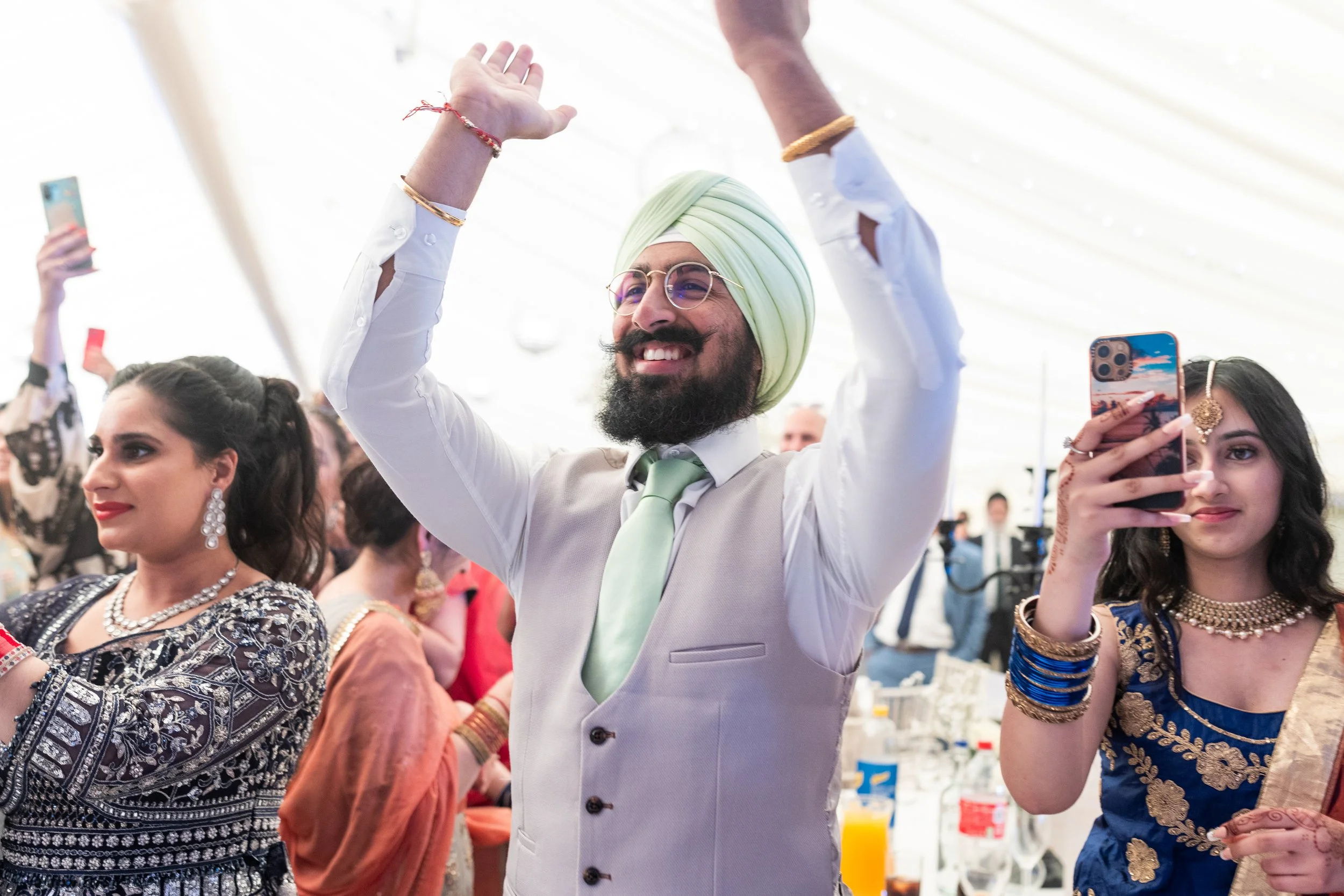 A group of people celebrating at a festive event, with a man in a light-colored vest, green tie, and turban smiling and raising his arms, while women around him are dressed in traditional Indian attire, with some taking photos.