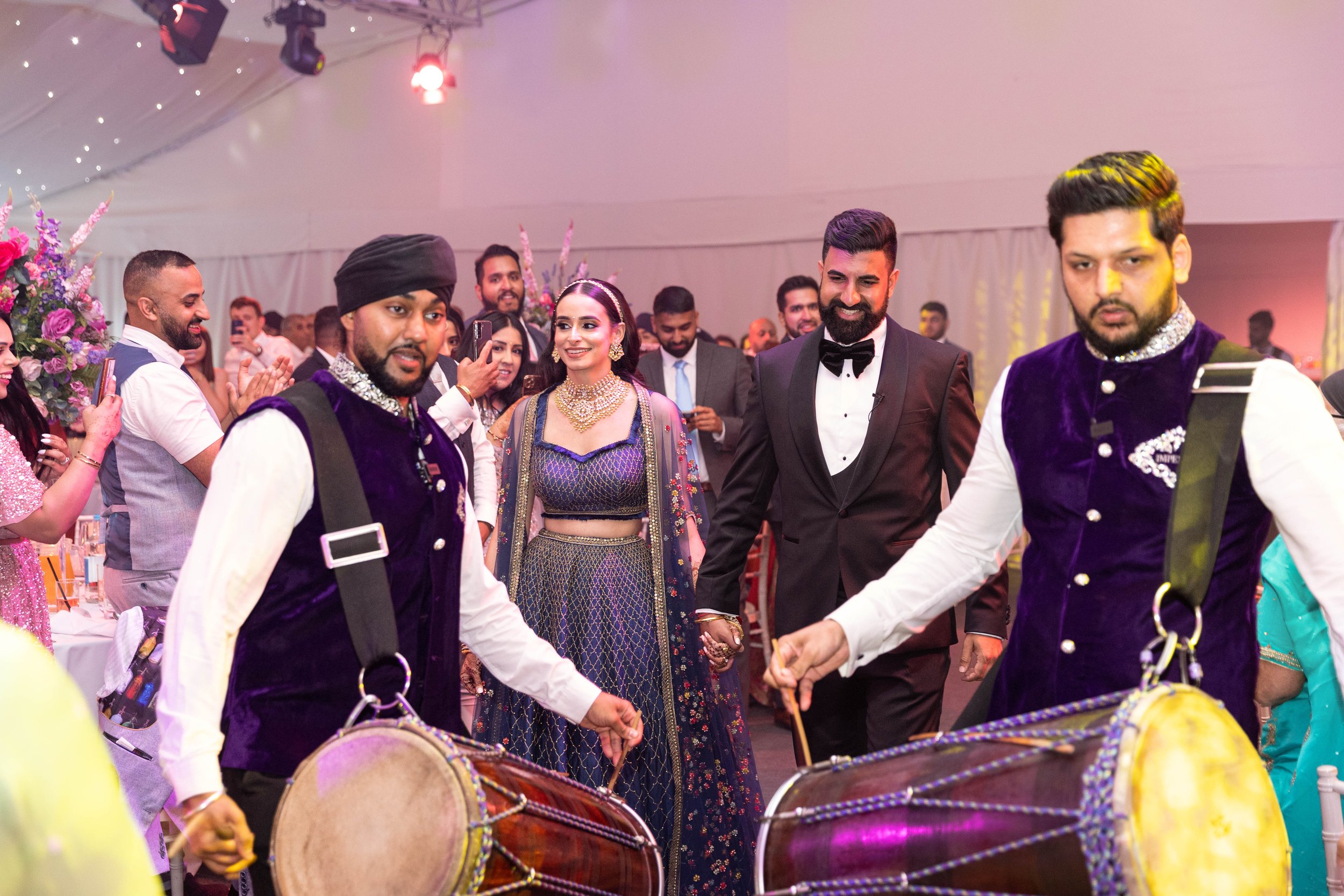 A group of people celebrating at a wedding reception, with a woman in a traditional dress and diamond jewelry holding hands with a man in a black tuxedo, dancing among other guests and musicians playing drums.