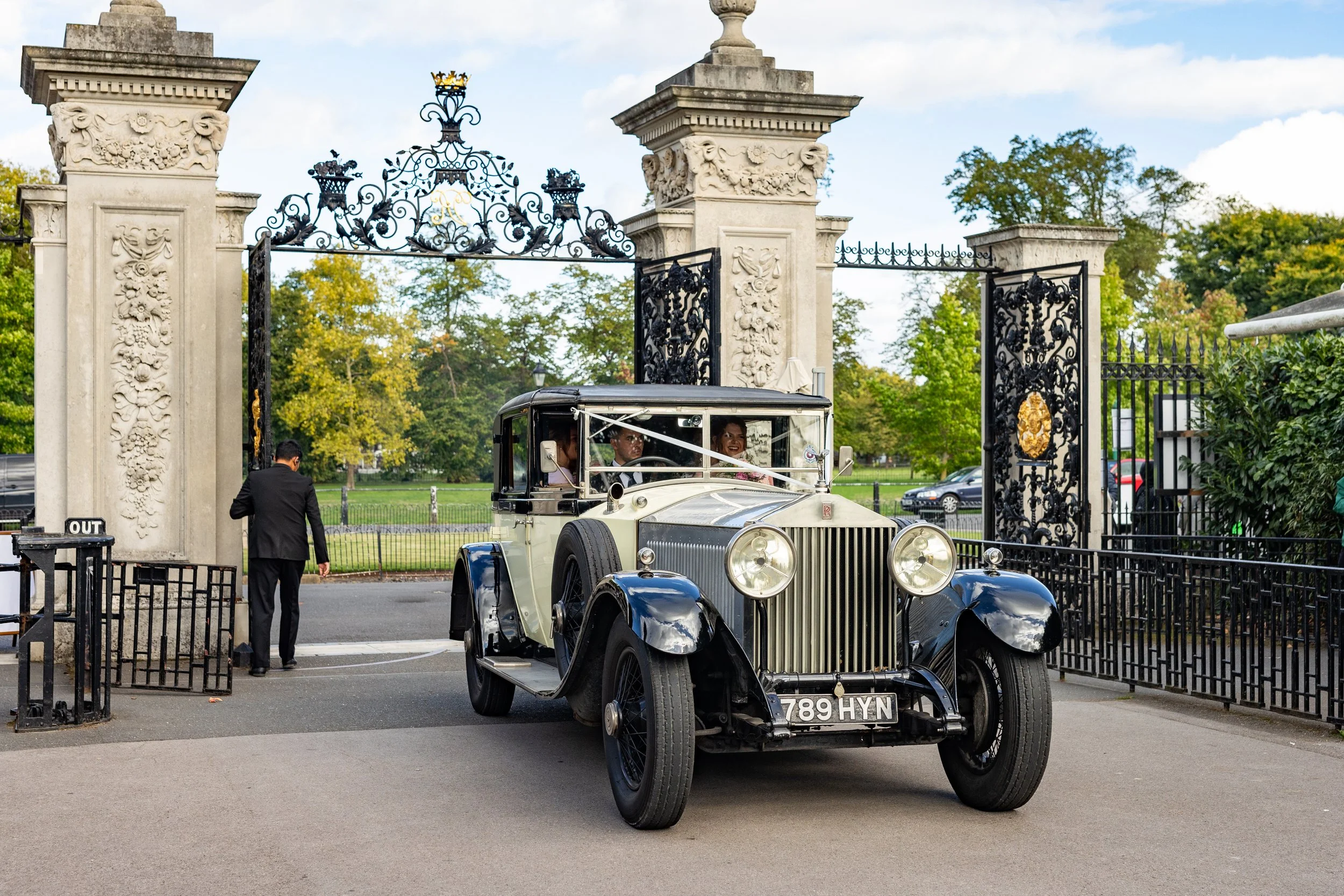 A vintage black and white car with round headlights and a license plate reading "789 HYN" driving through an ornate iron gate at the entrance of a park or estate. The background features greenery and a partly cloudy sky.