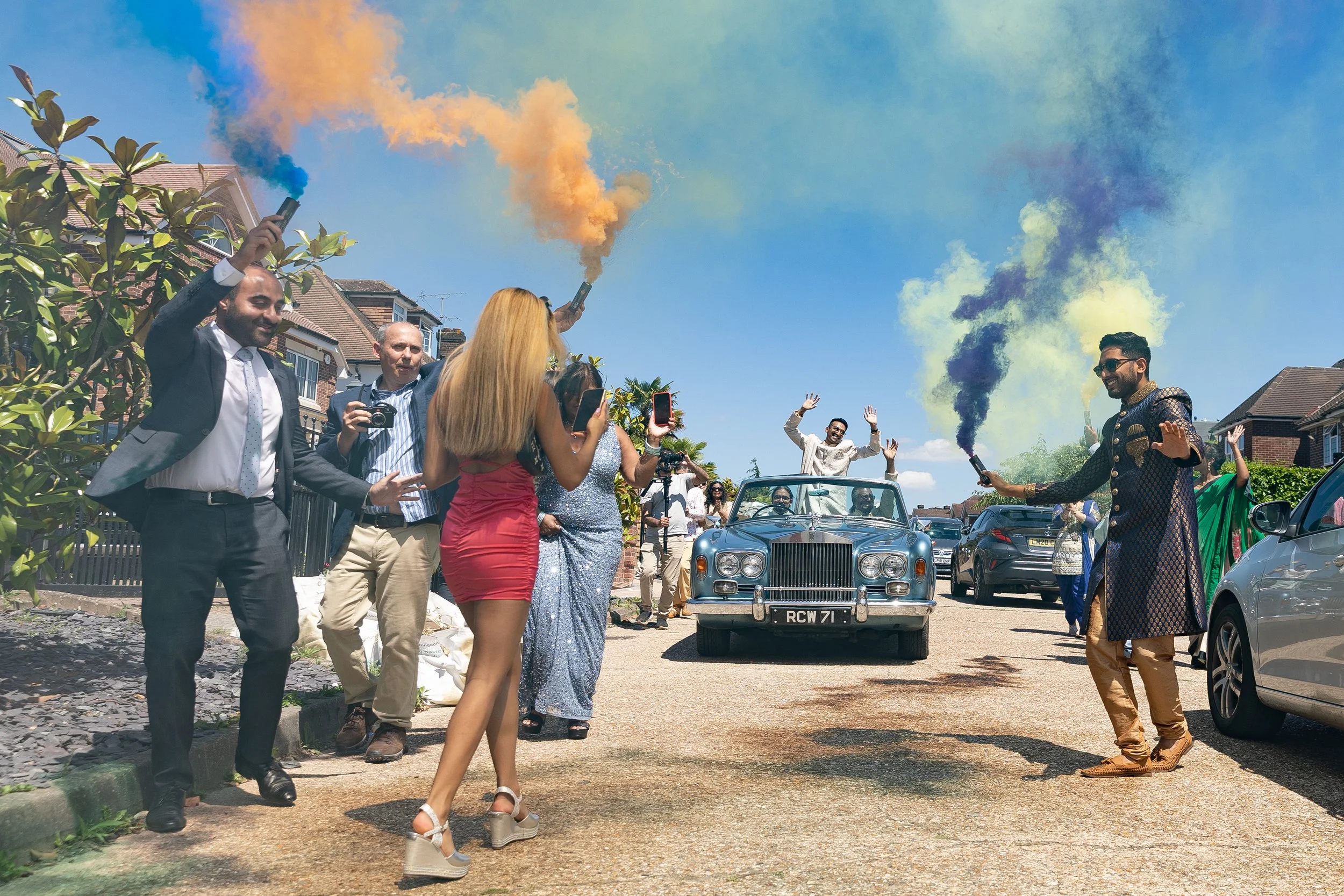 A group of people celebrating outdoors on a sunny day. There is a vintage car with a man standing through the sunroof waving. People are holding smoke bombs and taking photos, some with smartphones. The scene looks festive with colorful smoke in the sky.