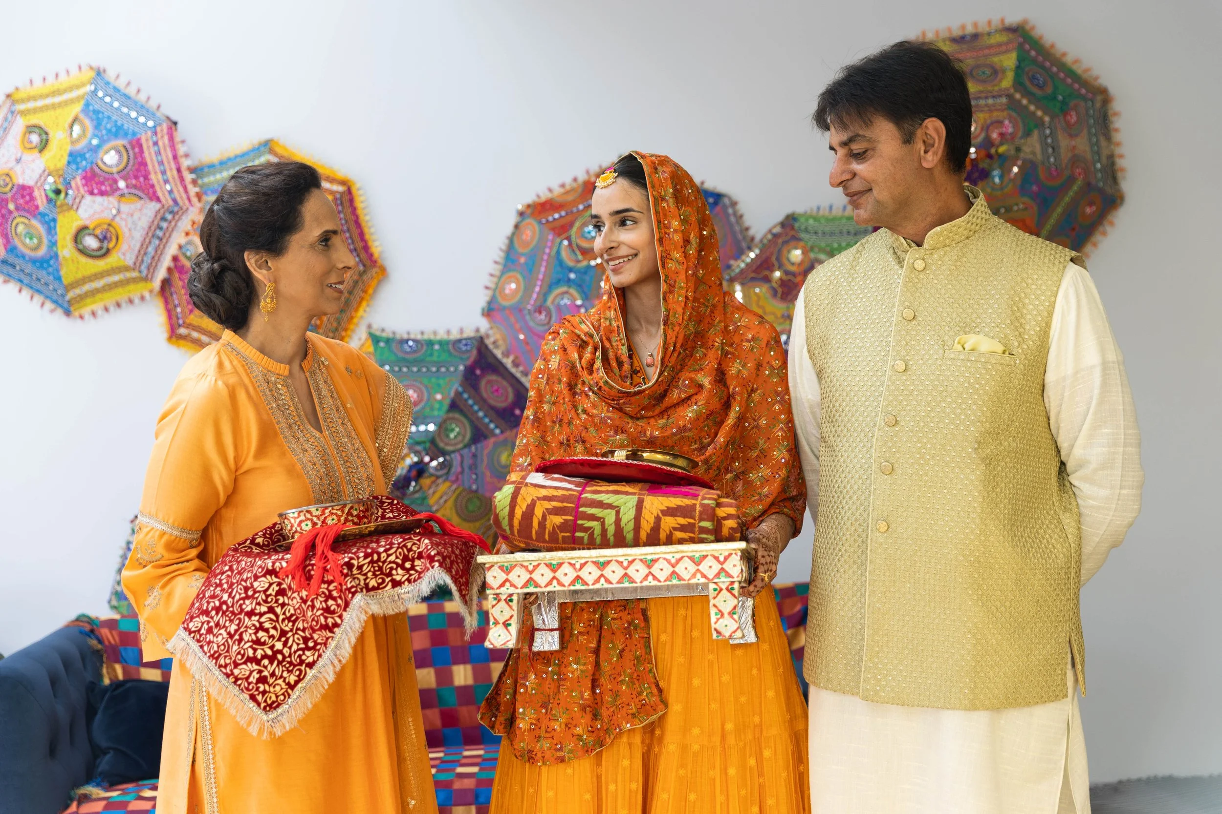A woman in orange traditional Indian attire with gold jewelry presents a gift to a smiling woman dressed in an orange and yellow saree with a matching headscarf, while a man in a cream and yellow outfit looks on. Colorful decorative umbrellas decorat