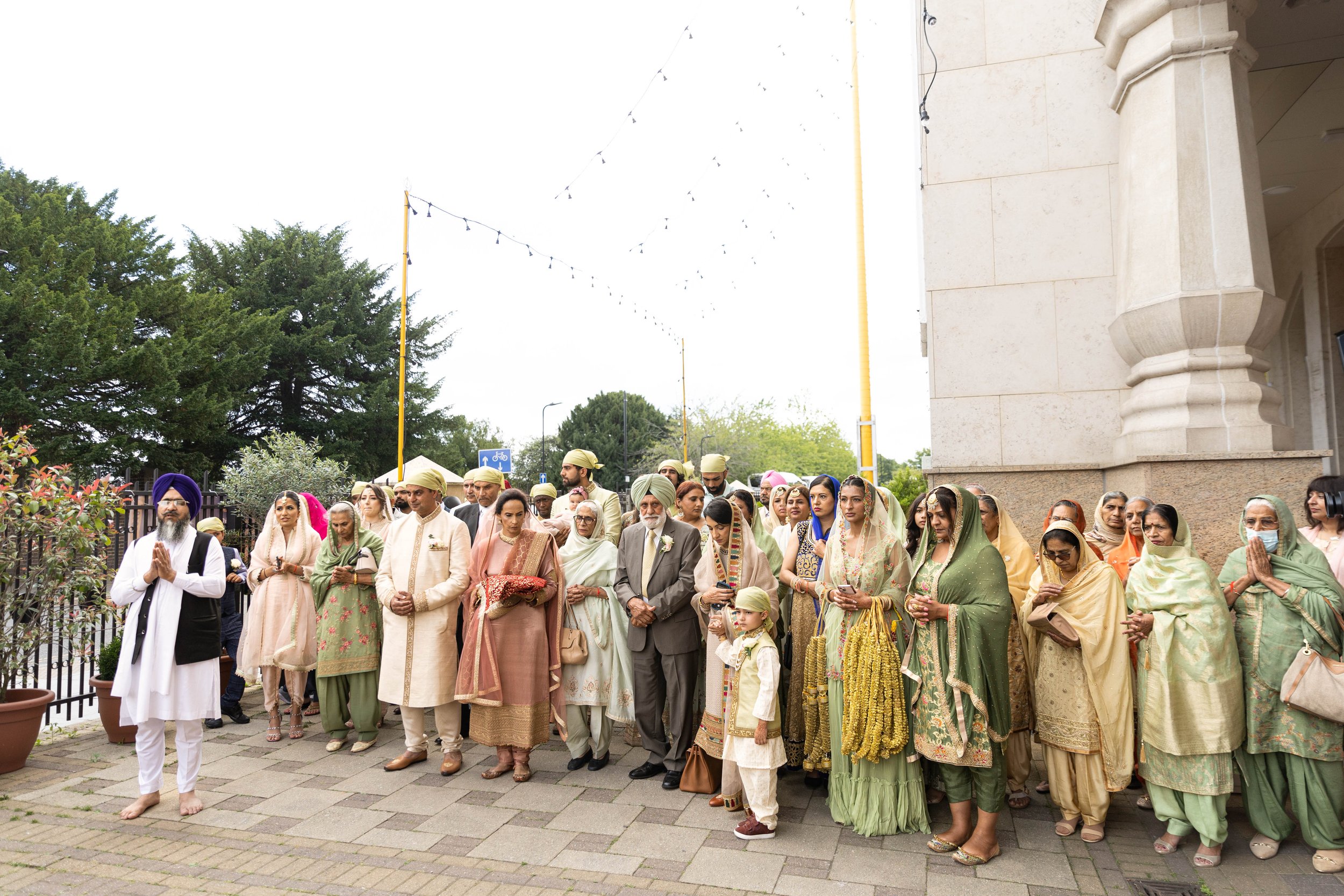 Group of Indian people dressed in traditional attire, standing outdoors near a building, possibly participating in a cultural or religious ceremony.