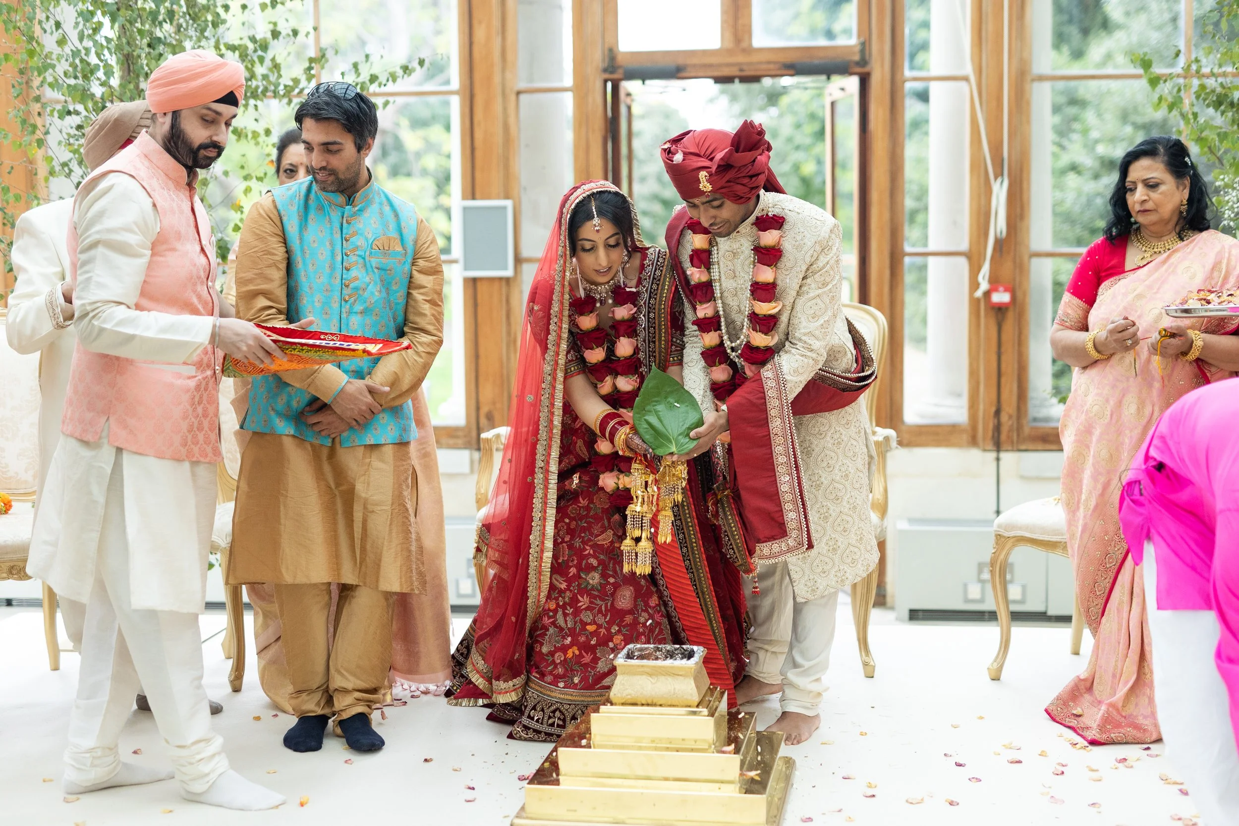 Indian wedding ceremony with bride and groom performing a ritual, surrounded by family members in traditional attire, indoors with large windows and greenery outside.