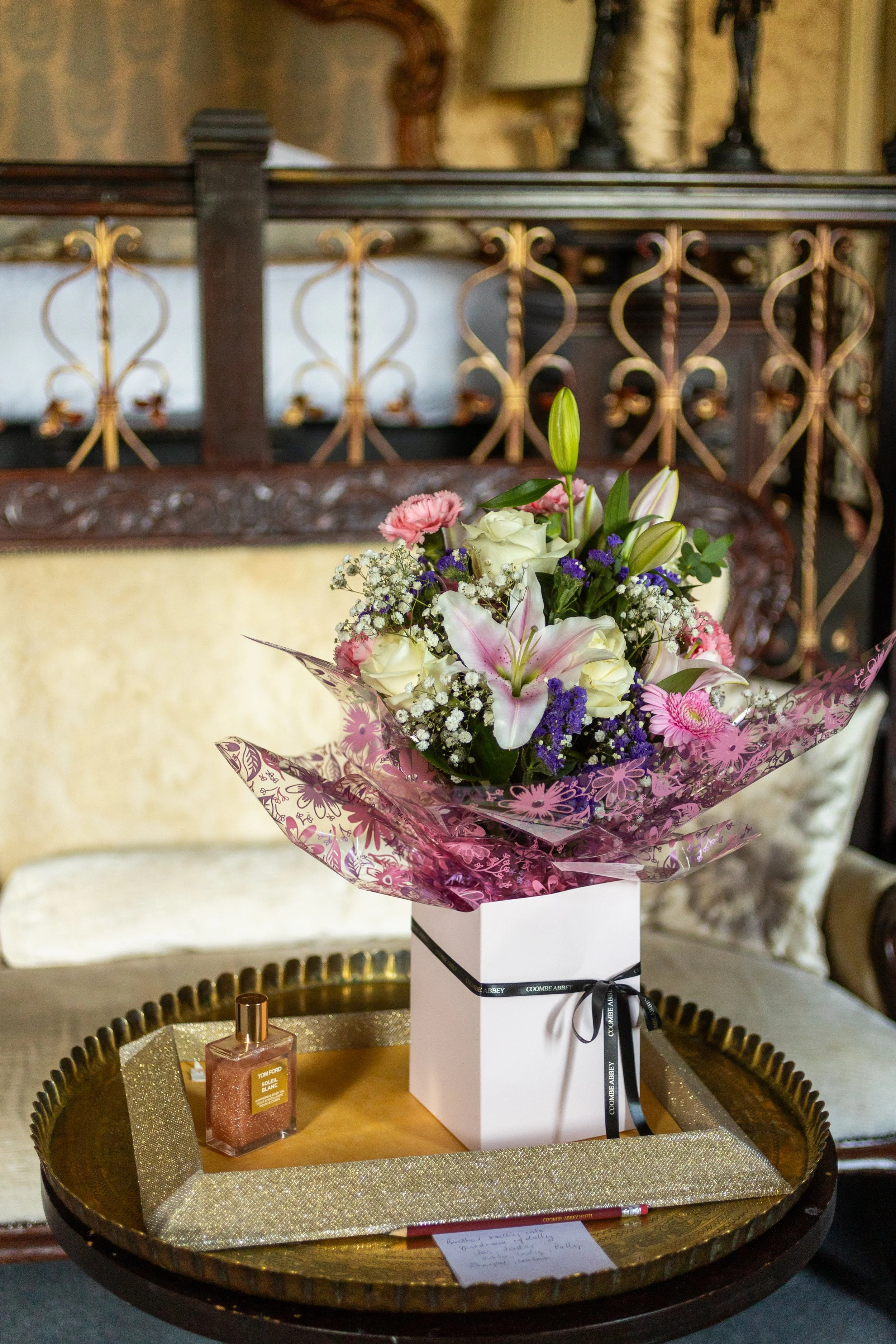 A bouquet of pink, white, purple, and green flowers in a pink and transparent wrap, placed on a white box with a black ribbon, on a gold tray with a small perfume bottle, pen, and notes in front of a vintage sofa with ornate wood and gold accents.