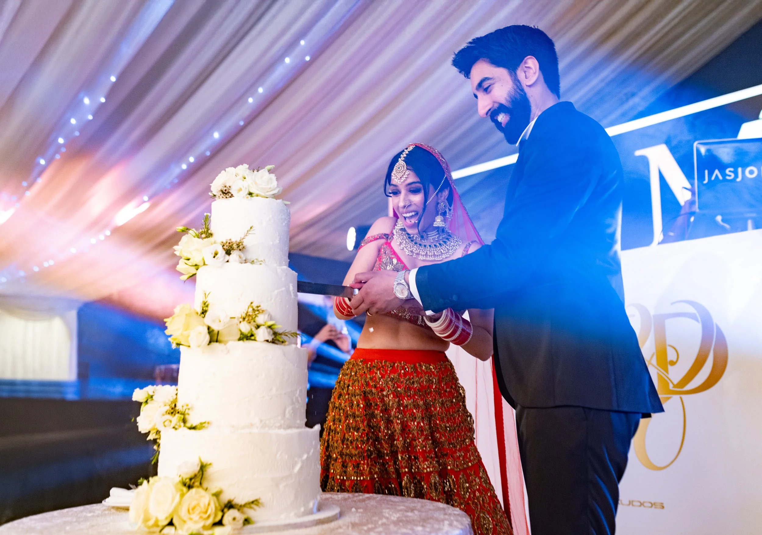 A bride and groom cutting a wedding cake together during their reception, wedding cake decorated with white flowers, bride wearing traditional Indian attire with jewelry, groom in a black suit, festive lighting and drapery in the background.