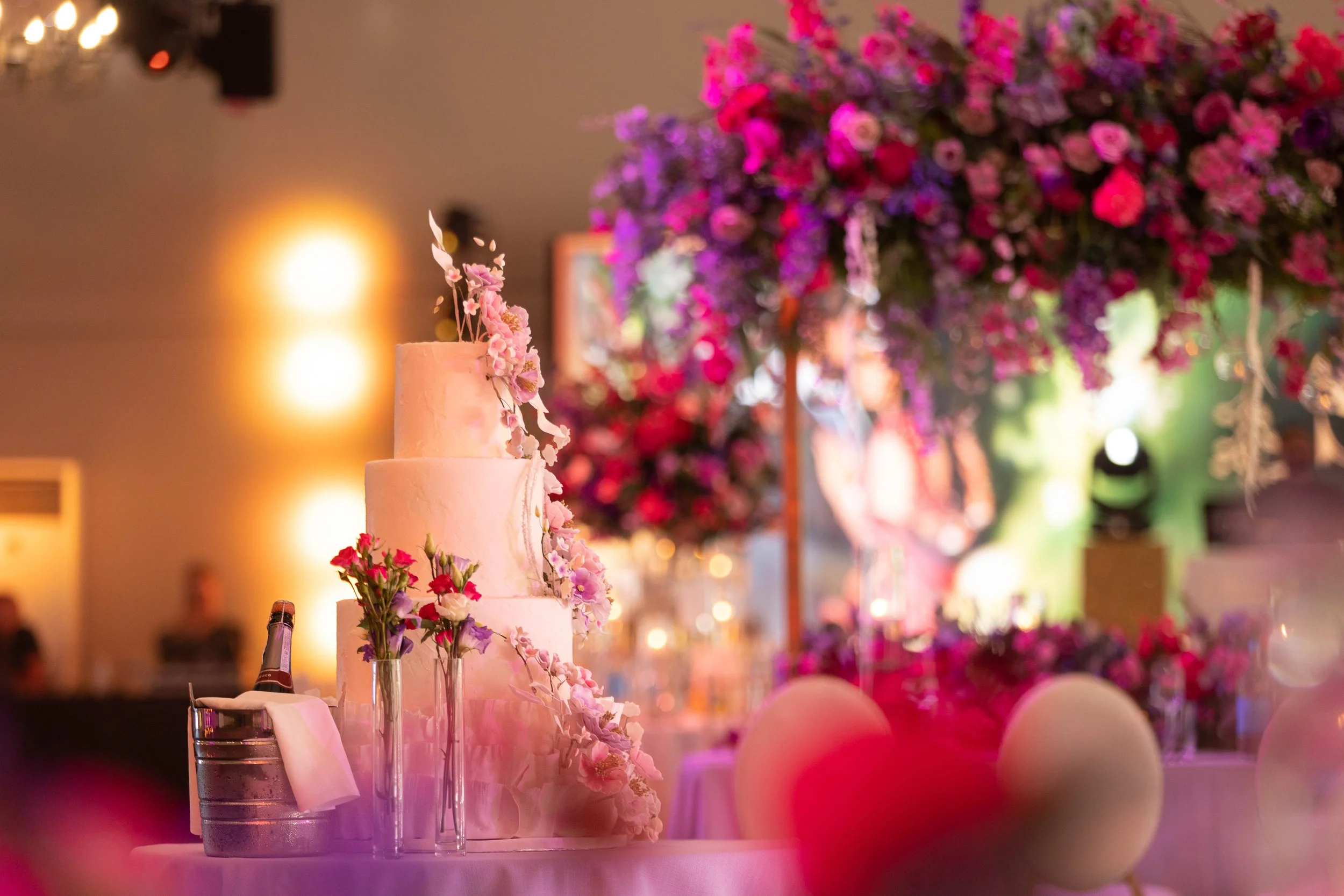 A wedding cake decorated with pink and purple flowers on a table with champagne bottles, surrounded by pink and white balloons at a decorated venue.