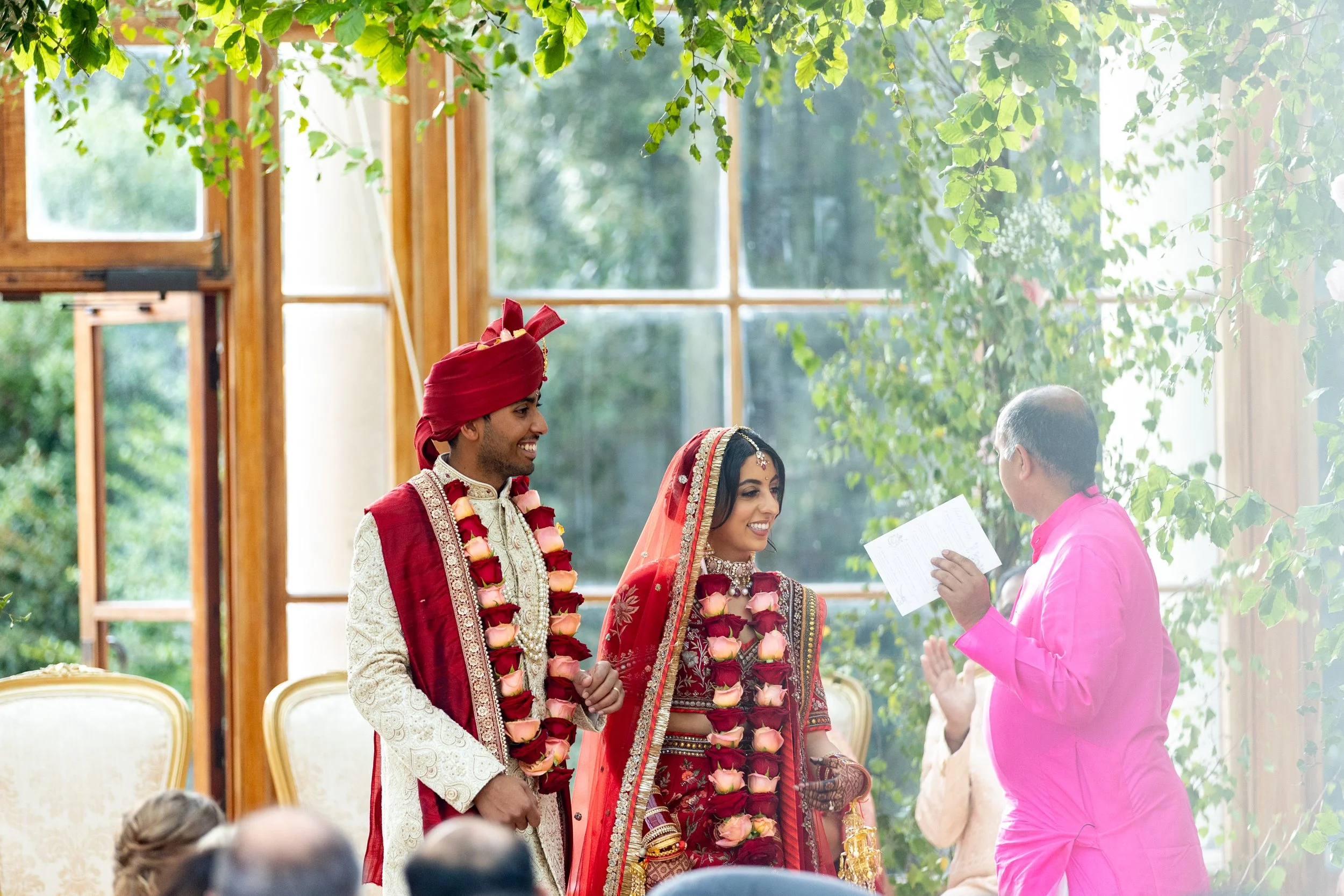 Indian wedding ceremony with bride and groom in traditional attire, wearing floral garlands, standing before officiant in a decorated indoor space with large windows and green foliage outside.