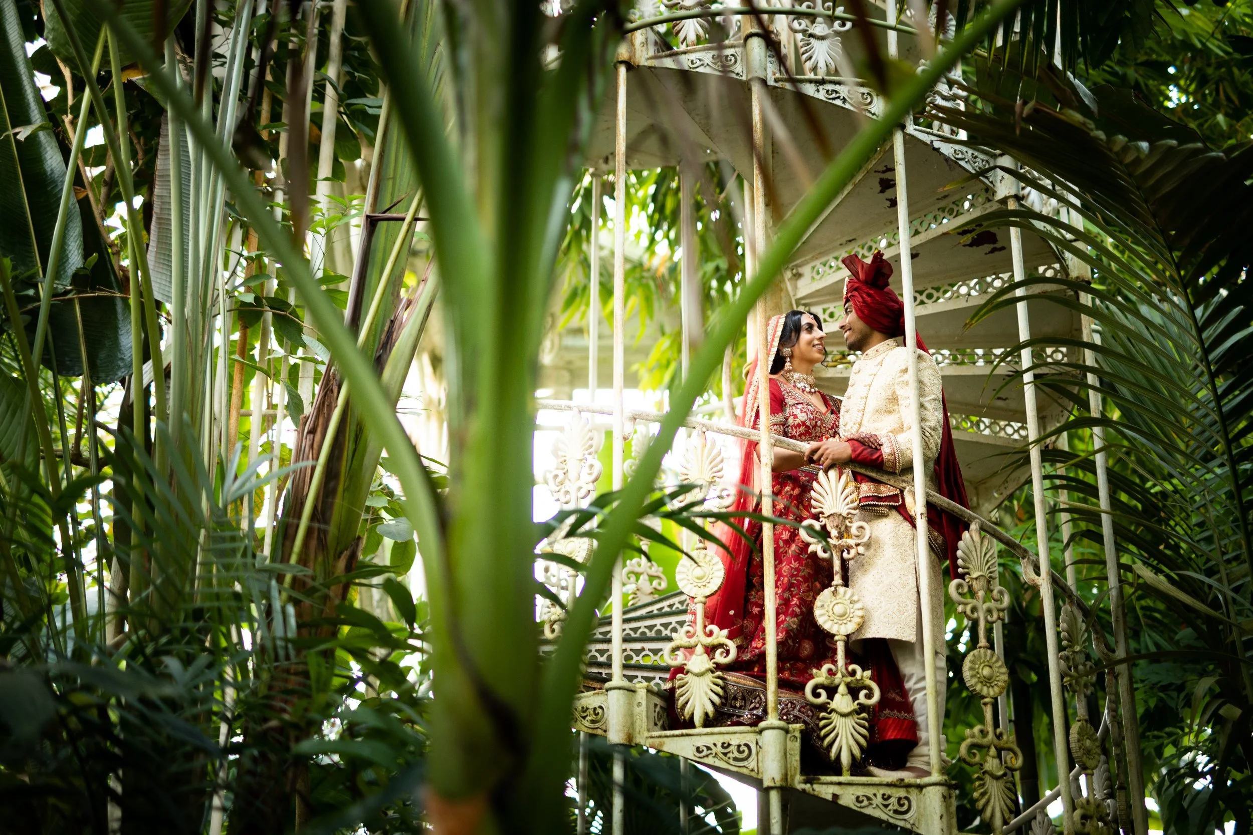 A couple in traditional Indian wedding attire standing on a decorative white metal staircase surrounded by lush green plants and trees in a jungle setting, gazing at each other affectionately.