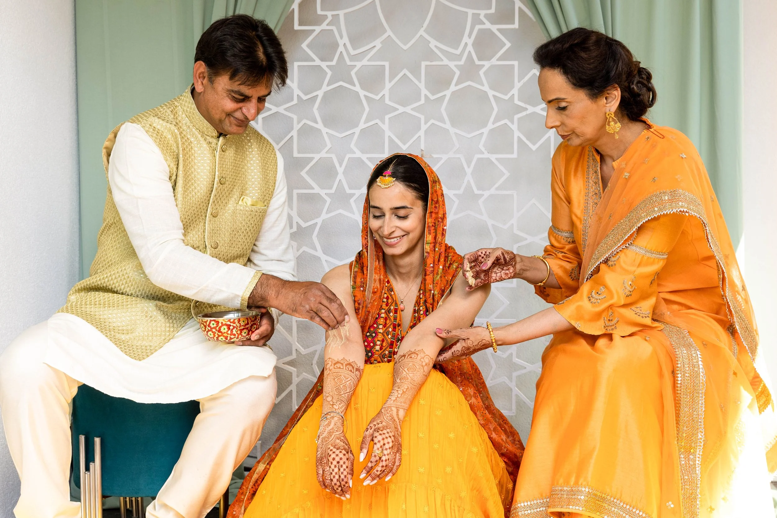 A woman in traditional Indian wedding attire is being decorated with mehndi by two women, one on each side, while a man sits beside her holding a decorative bowl.