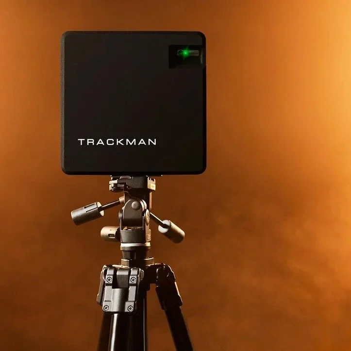 A black square device labeled 'TRACKMAN' mounted on a tripod against a brown background, with a green light indicator on the top right corner of the device.