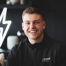 Smiling young man with short hair, wearing a black jacket, standing in front of a dark background with a white lightning bolt design.