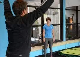 A person taking a photo of a woman standing inside a gym near large windows.