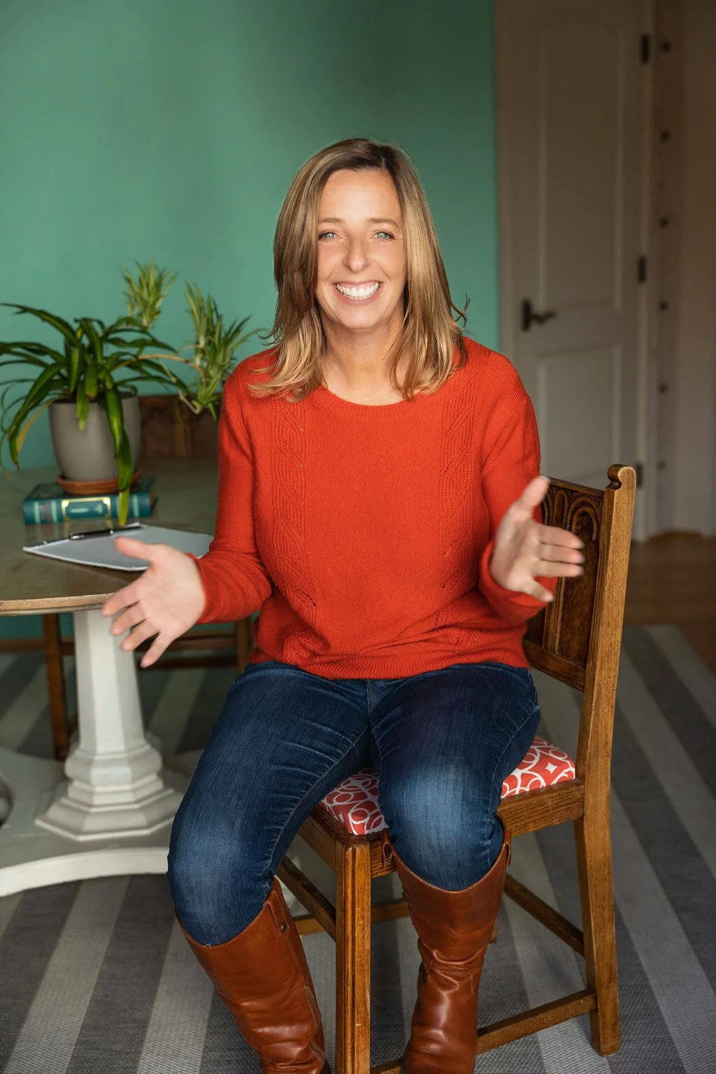 Woman in red sweater sitting on chair in room with teal walls, holding a tablet or paper, and smiling.
