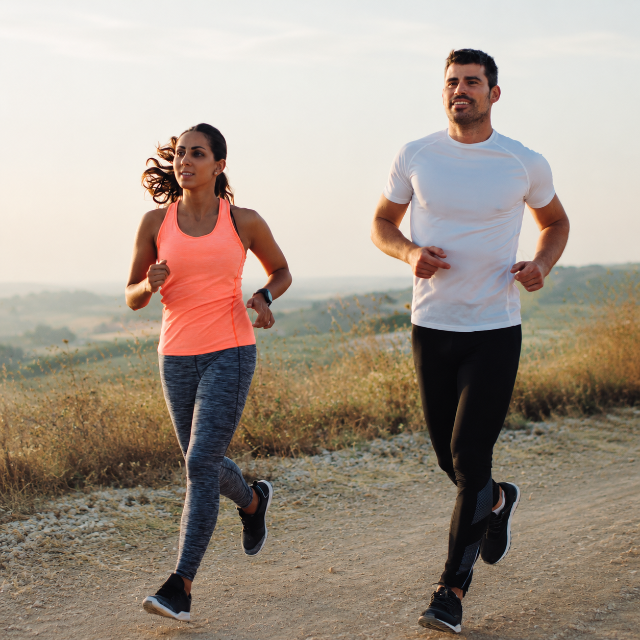 A man and woman jogging outdoors on a dirt trail during daytime with a hazy background of hills and sky.