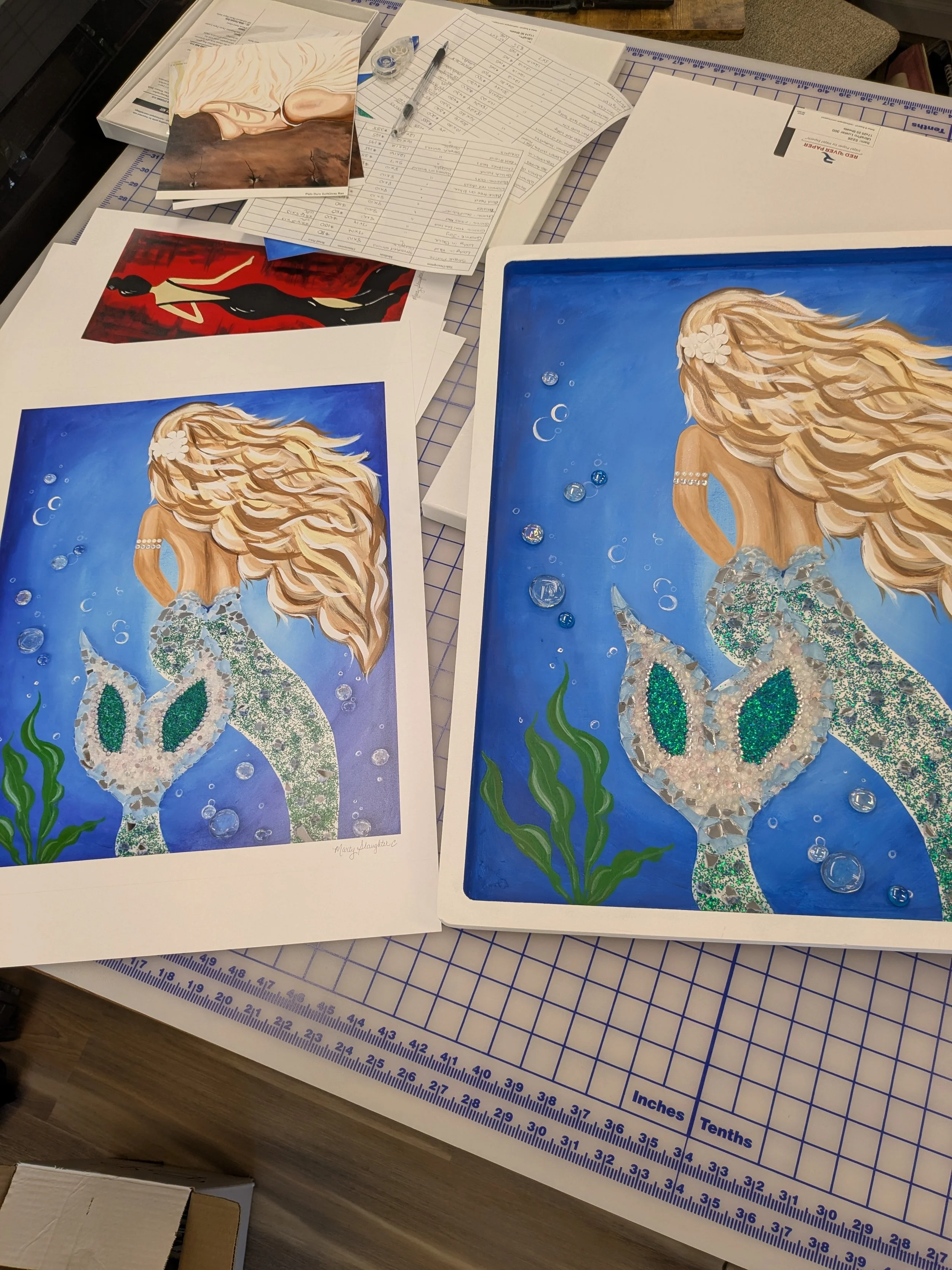 A work table with art prints, sketches, and a reference photo of a mermaid painting, with a grid-lined cutting mat underneath.