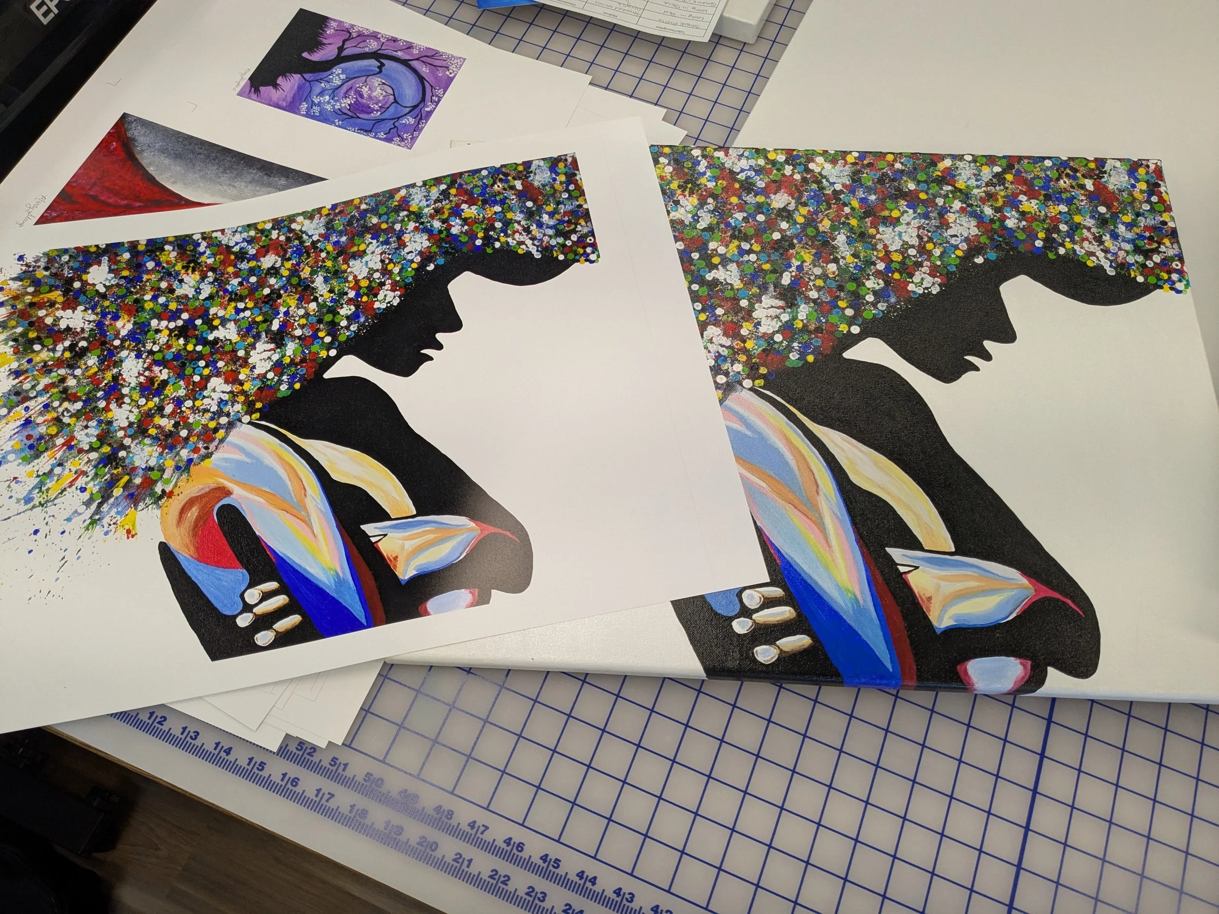 Multiple printed artworks featuring silhouettes of a woman's profile with colorful, abstract, and pointillist styles, placed on a workspace table with grid cutting mat and paperwork.