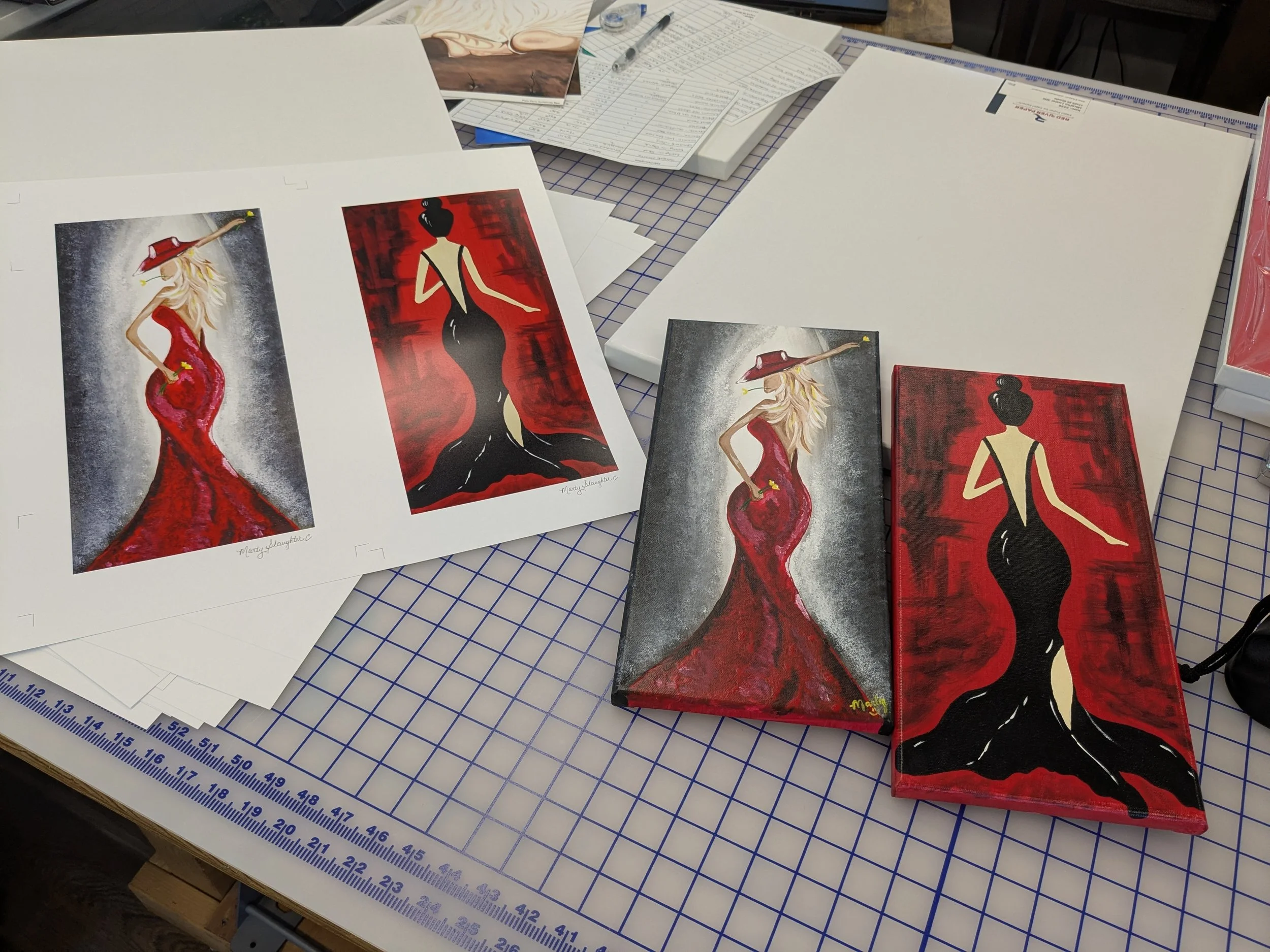 Printouts and artwork featuring fashion illustrations of women in elegant dresses on a worktable covered with a grid cutting mat, with papers, pens, and tools scattered around.