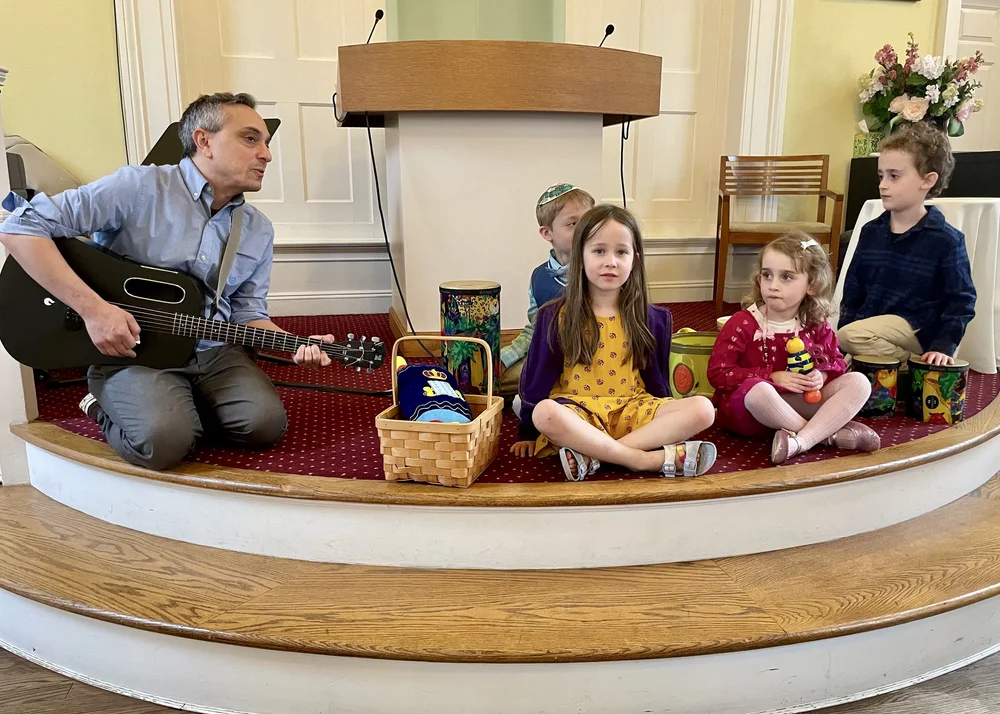 A rabbi playing guitar for children sitting in a circle on the bimah.